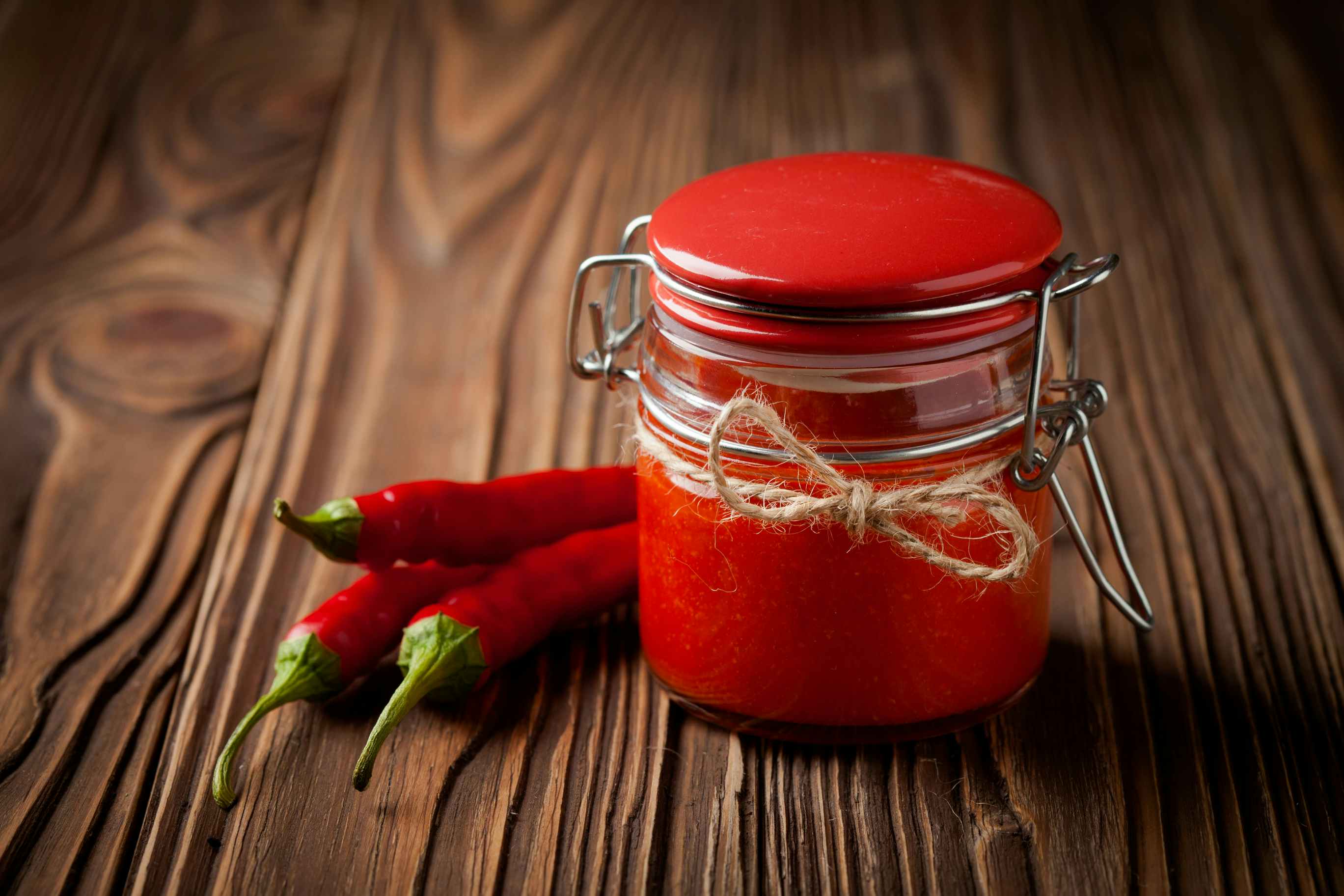 Jar of homemade sriracha sauce with chili peppers on a wooden table.