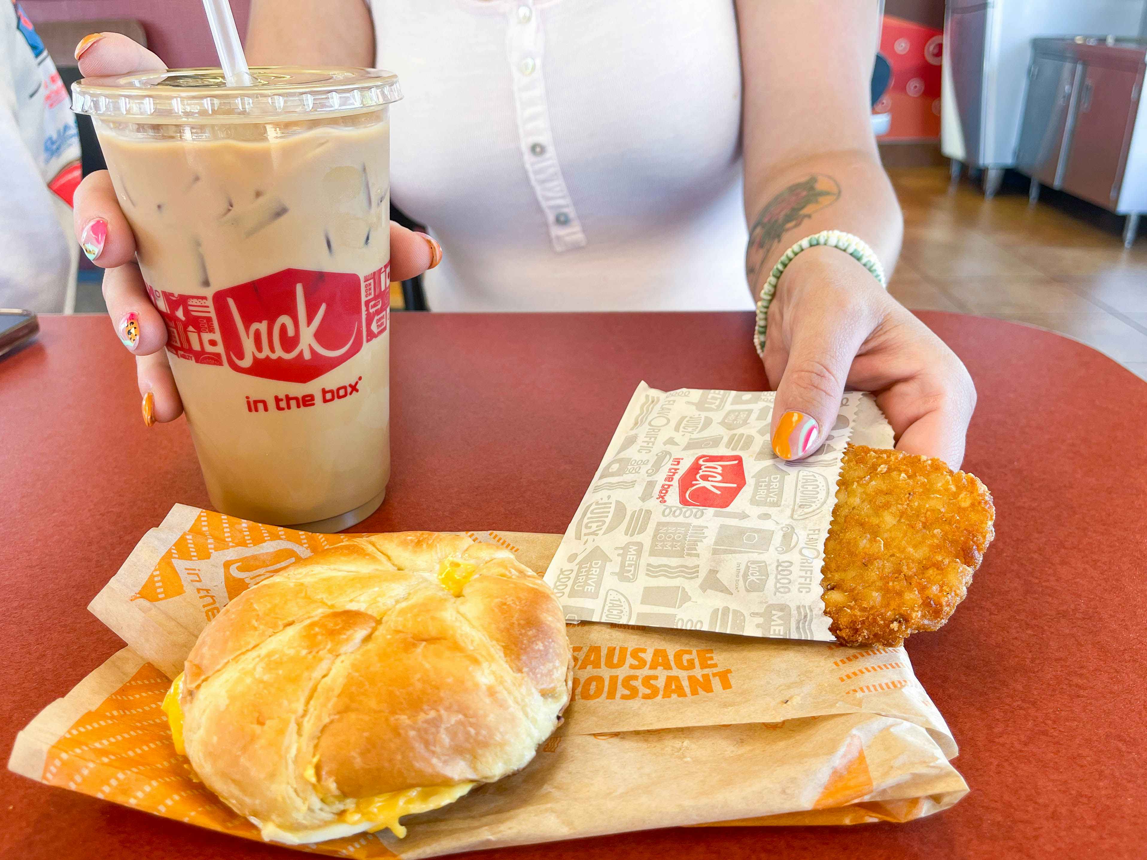 a person holding a iced coffee and hashbrown with jack in the box breakfast sandwich on table