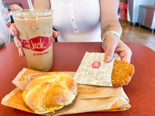 a person holding a iced coffee and hashbrown with jack in the box breakfast sandwich on table