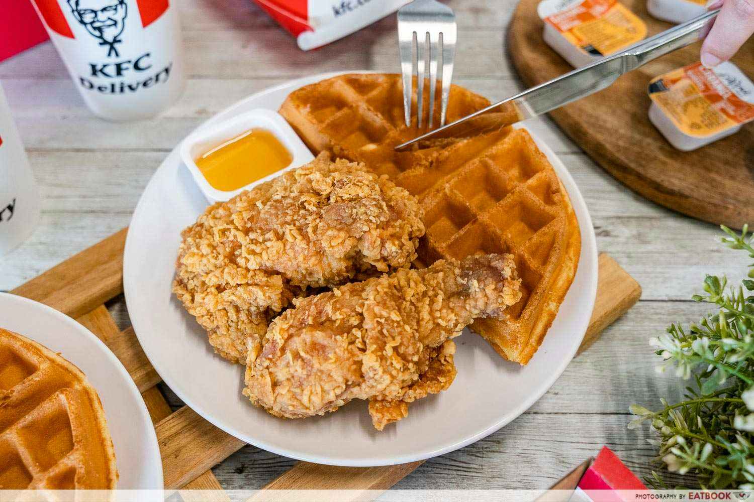 A plate of Chicken and waffles with KFC cup, and food container in the background.