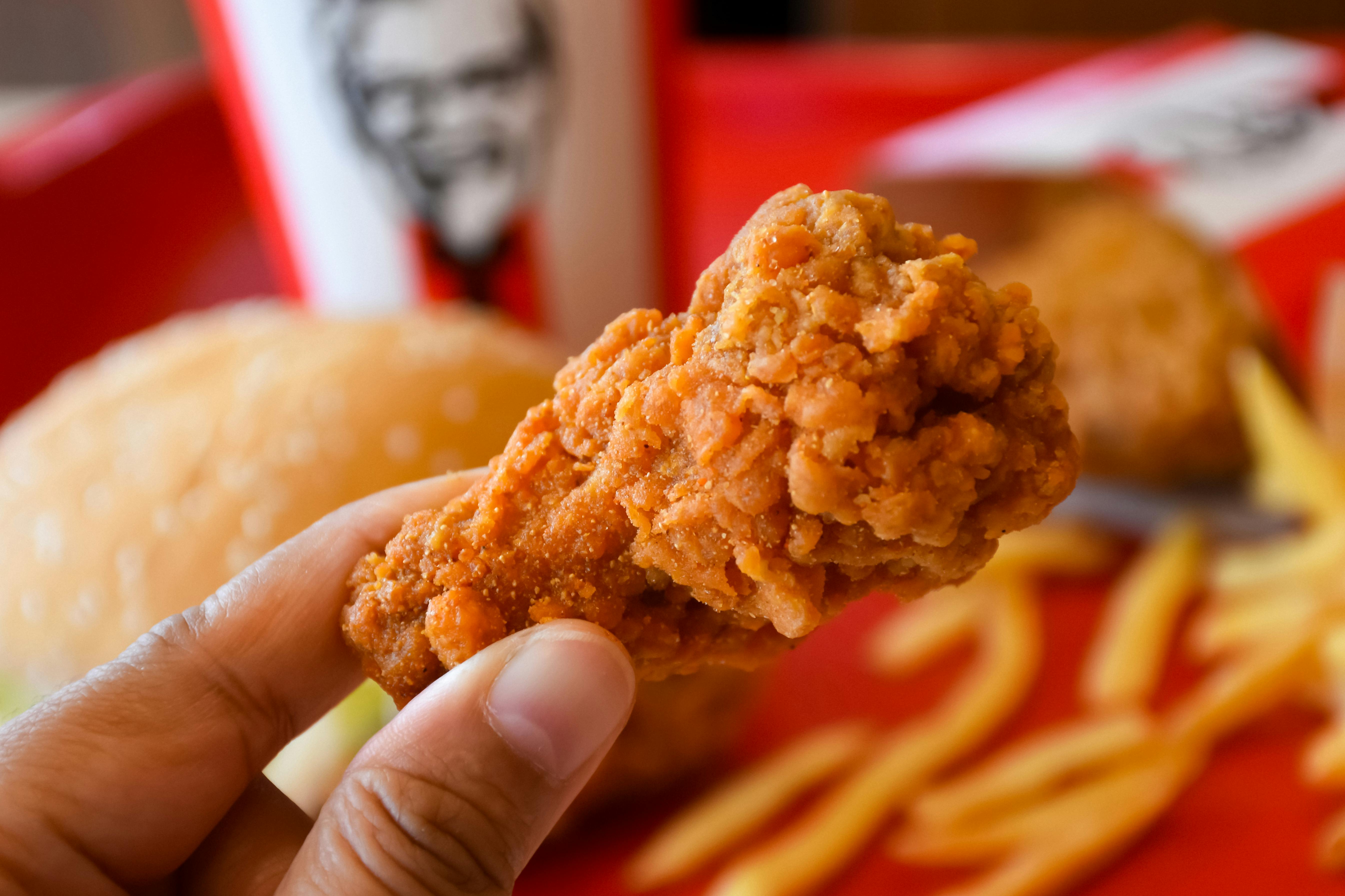 A close of a of chicken wing in a persons hand with other KFC food and a KFC drink cup in the background.