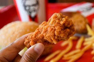 A close of a of chicken wing in a persons hand with other KFC food and a KFC drink cup in the background.