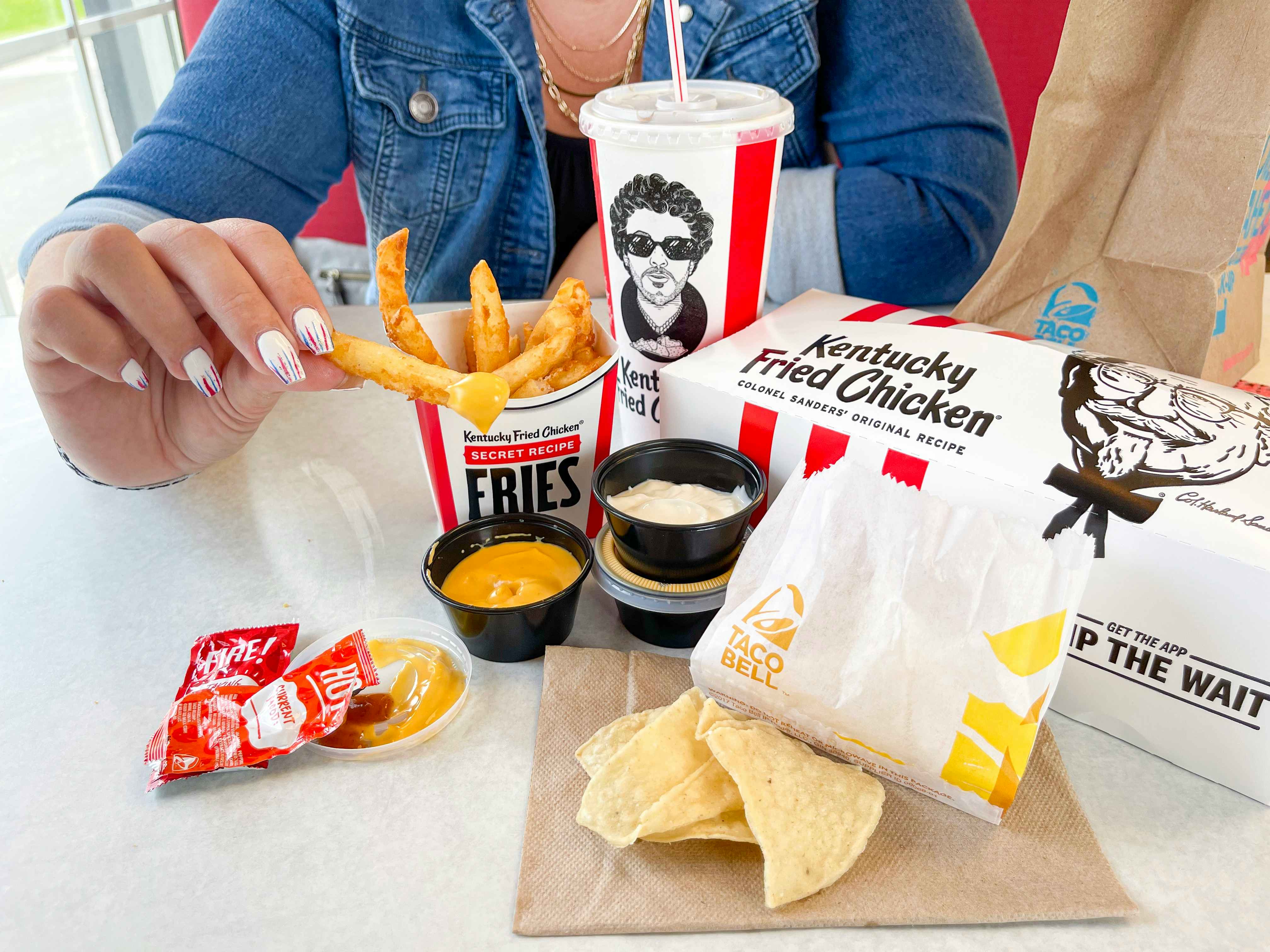 A KFC box of fries on a table next to a KFC box, a Taco Bell paper bag, some chips, nacho cheese sauce, and Taco Bell hot and fire sauce packets with a person holding a fry that has been dipped in nacho cheese..
