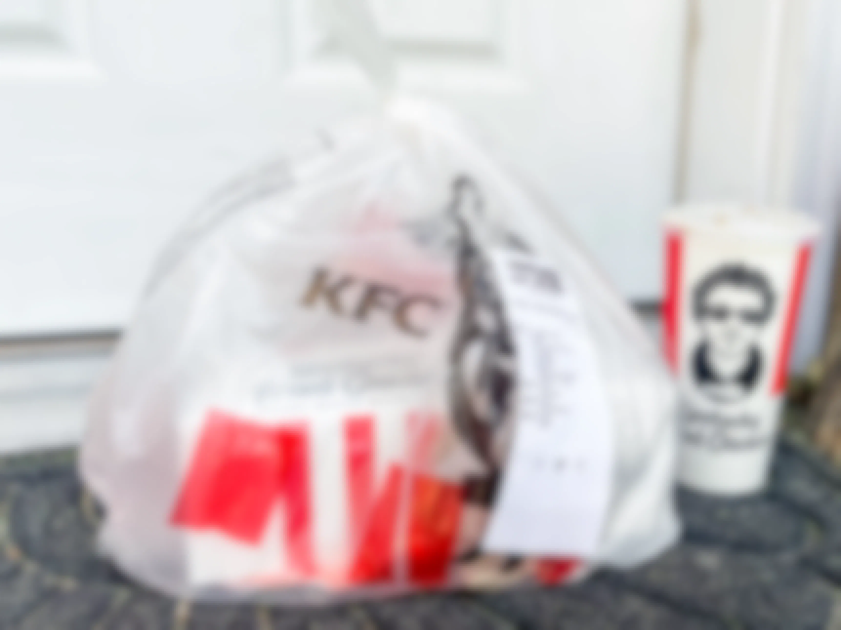 A bag of KFC takeout sitting on a front porch next to a drink.