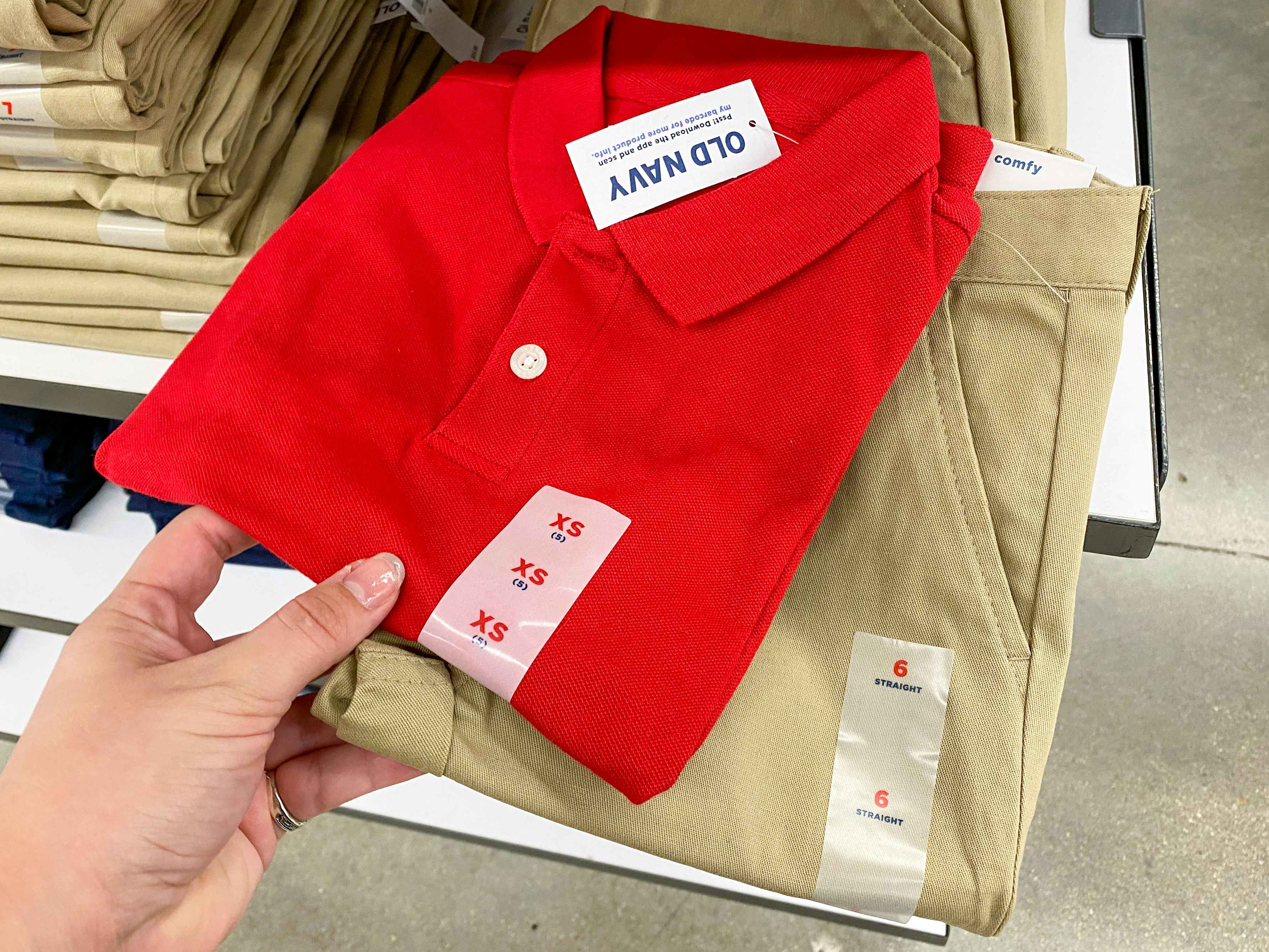 A person's hand taking a children's school uniform polo shirt and a pair of uniform pants from a shelf at Old Navy.