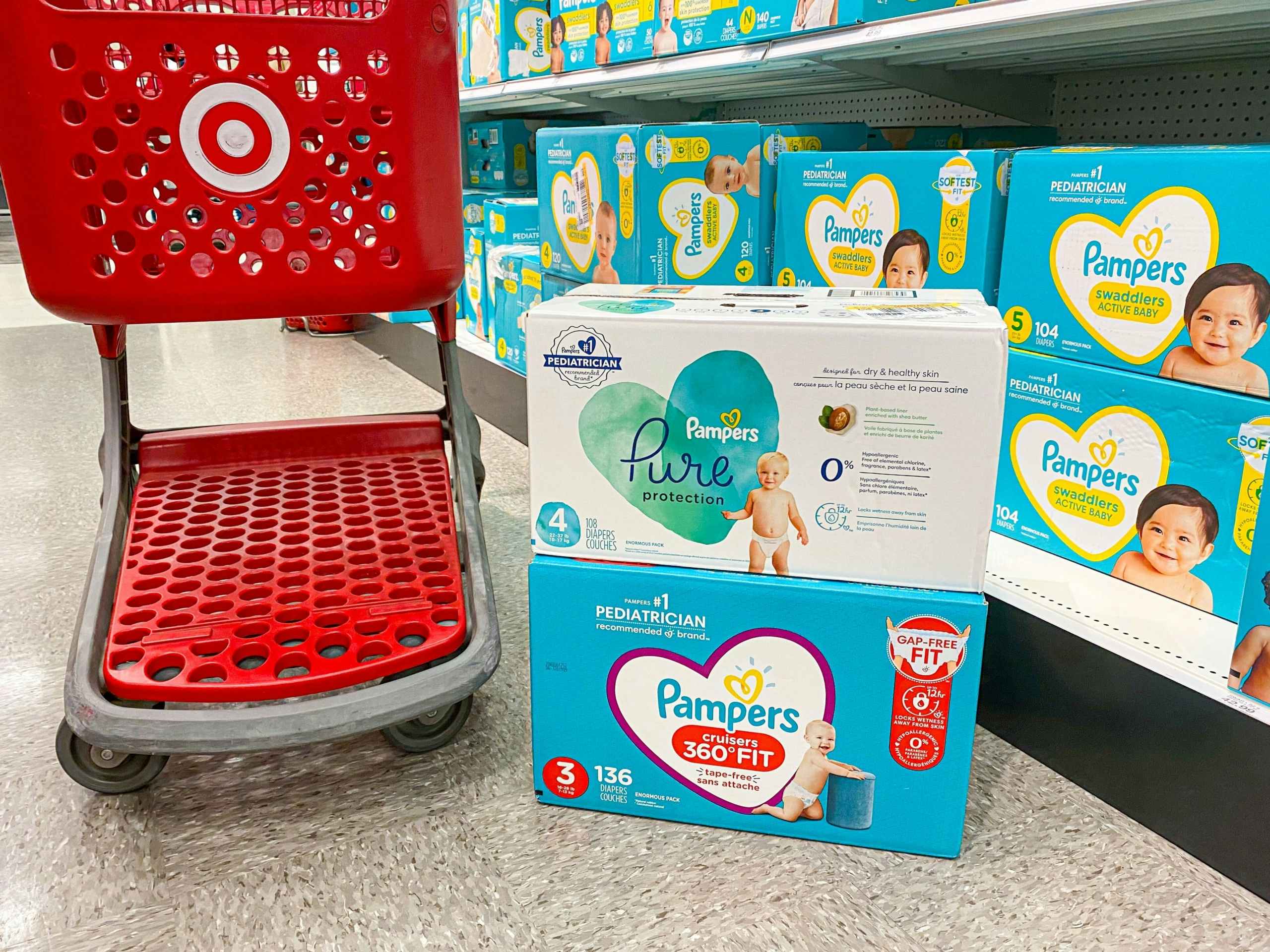 Two boxes of Pampers diapers stacked on the ground next to a Target shopping cart in the store aisle