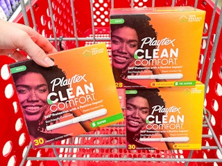 Hand holding up 1 box of Playtex tampons in front of a Target shopping cart that has two other boxes of Playtex tampons in it