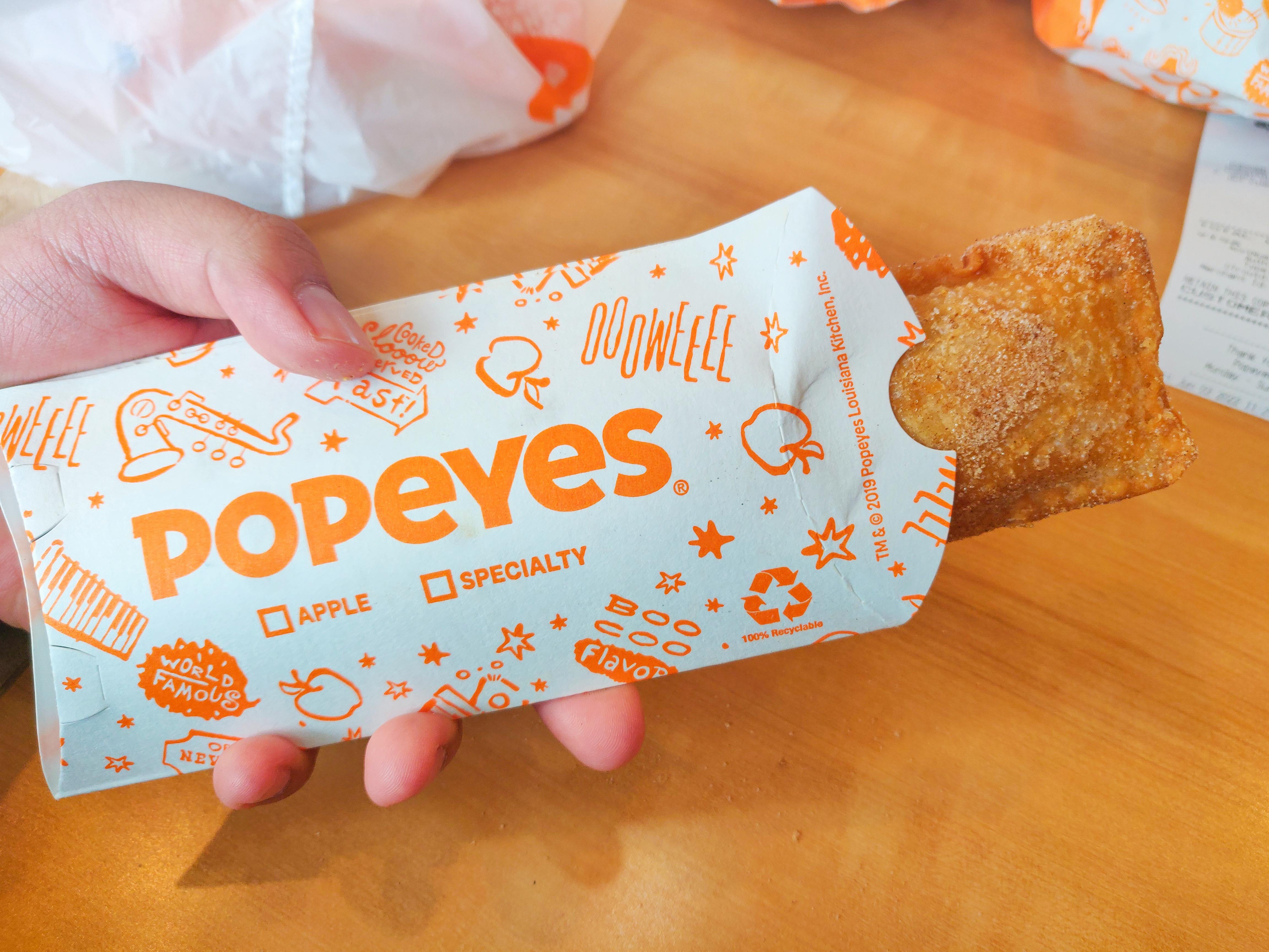 A person's hand holding a Popeyes apple pie treat above a table inside Popeyes.