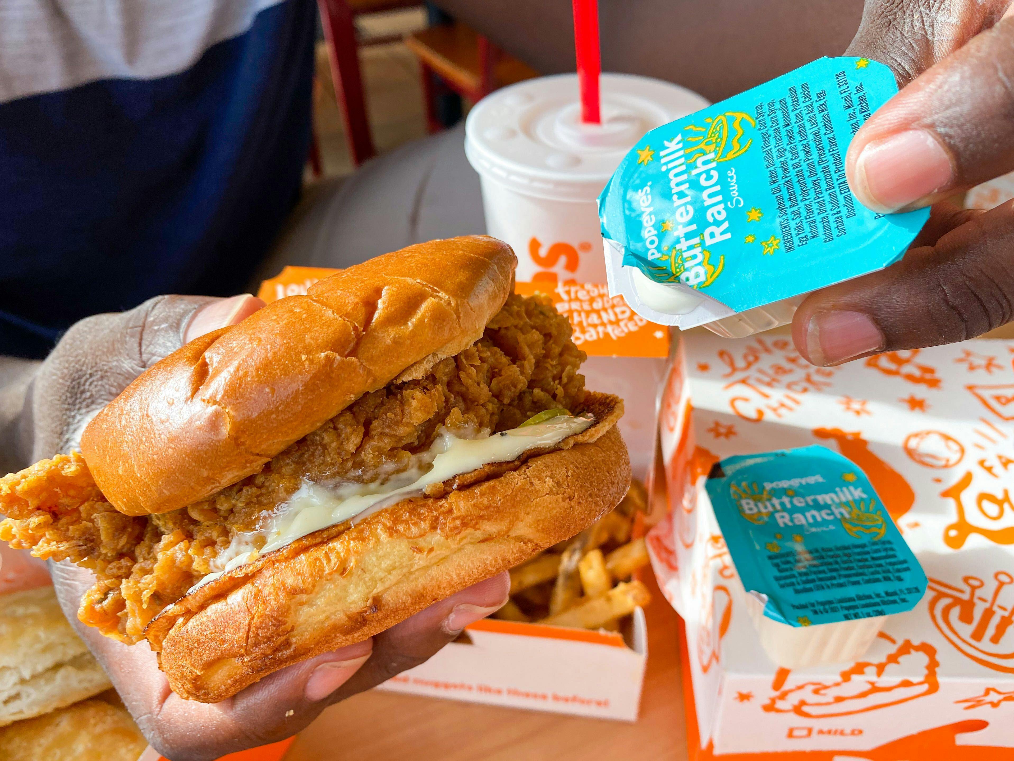 A person's hands holding a Popeyes chicken sandwich and a container of Buttermilk Ranch at a table in Popeyes.