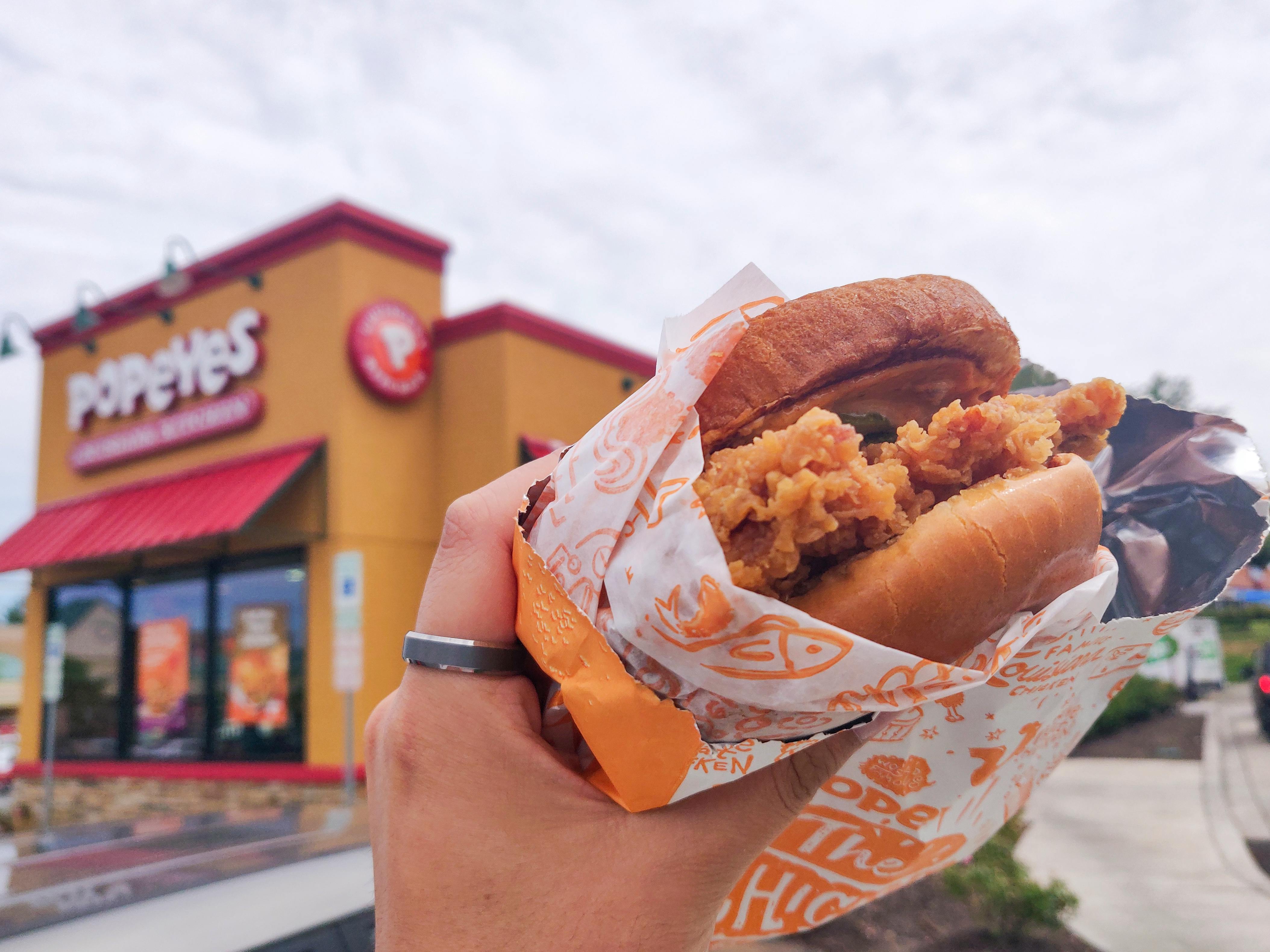 A person's hand holding a Popeyes chicken sandwich with a Popeyes restaurant in the background.