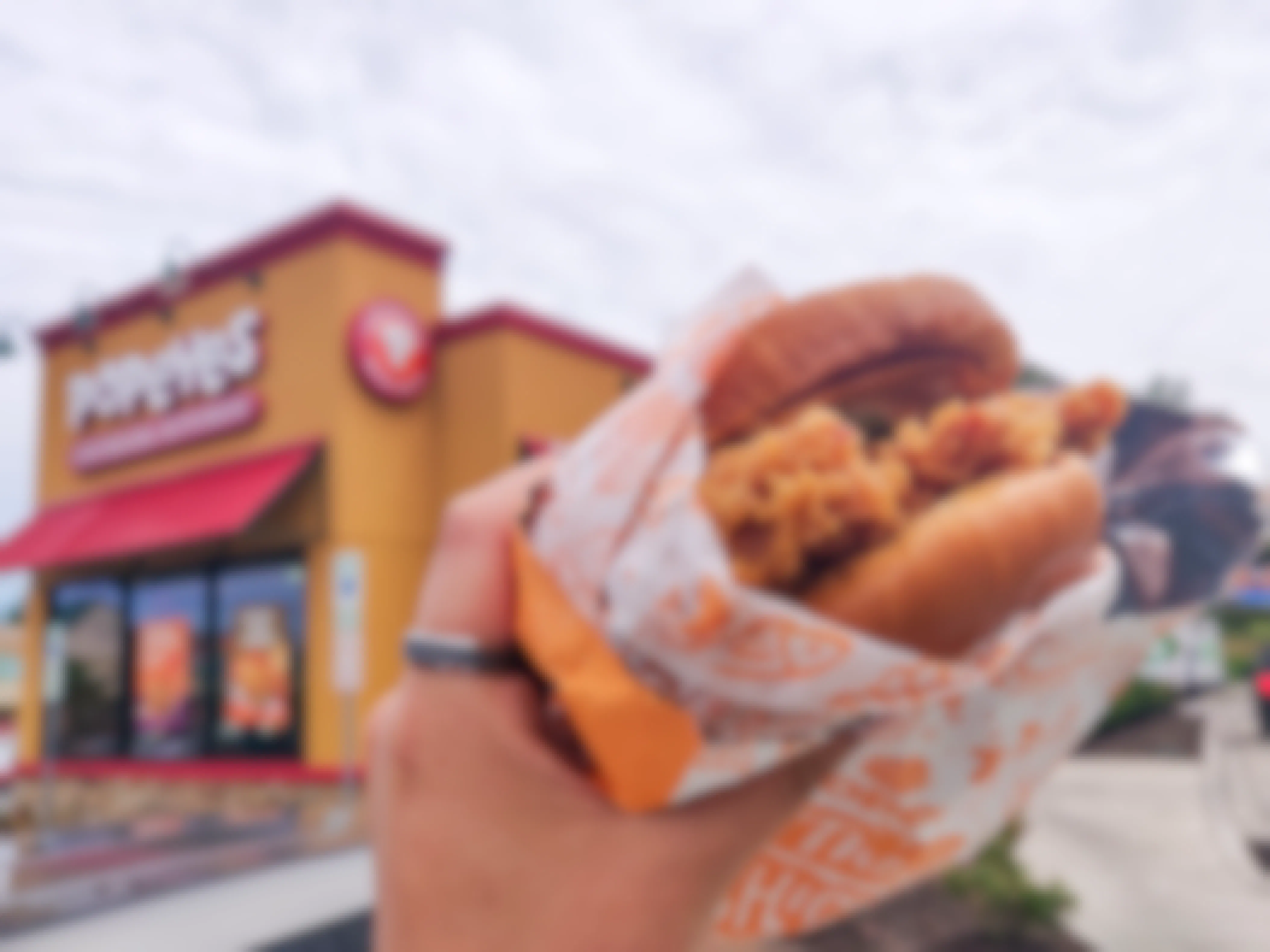 A person's hand holding a Popeyes chicken sandwich with a Popeyes restaurant in the background.