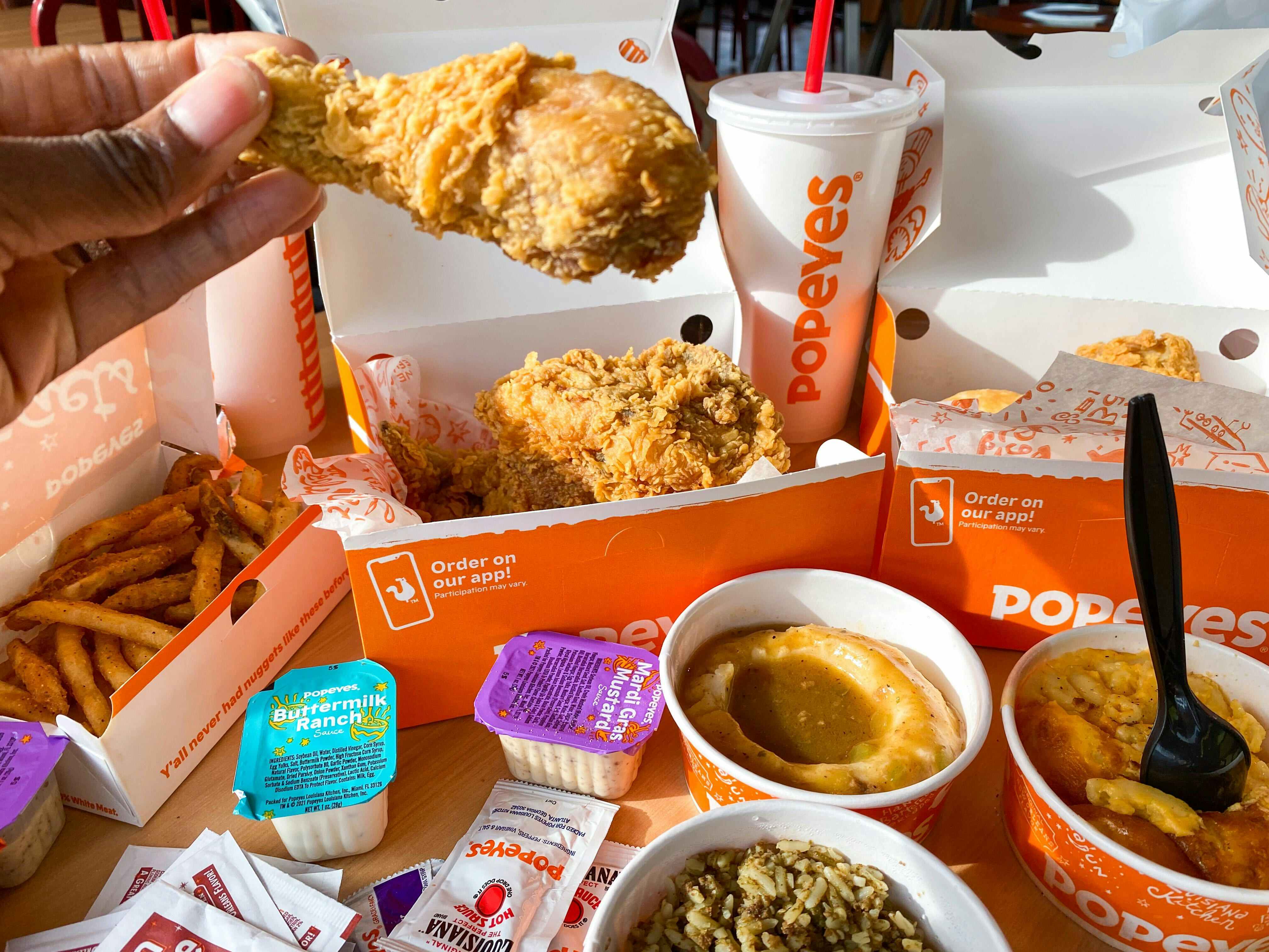 A person's hand holding up a piece of chicken above a Popeyes chicken family meal displayed on a table at Popeyes.