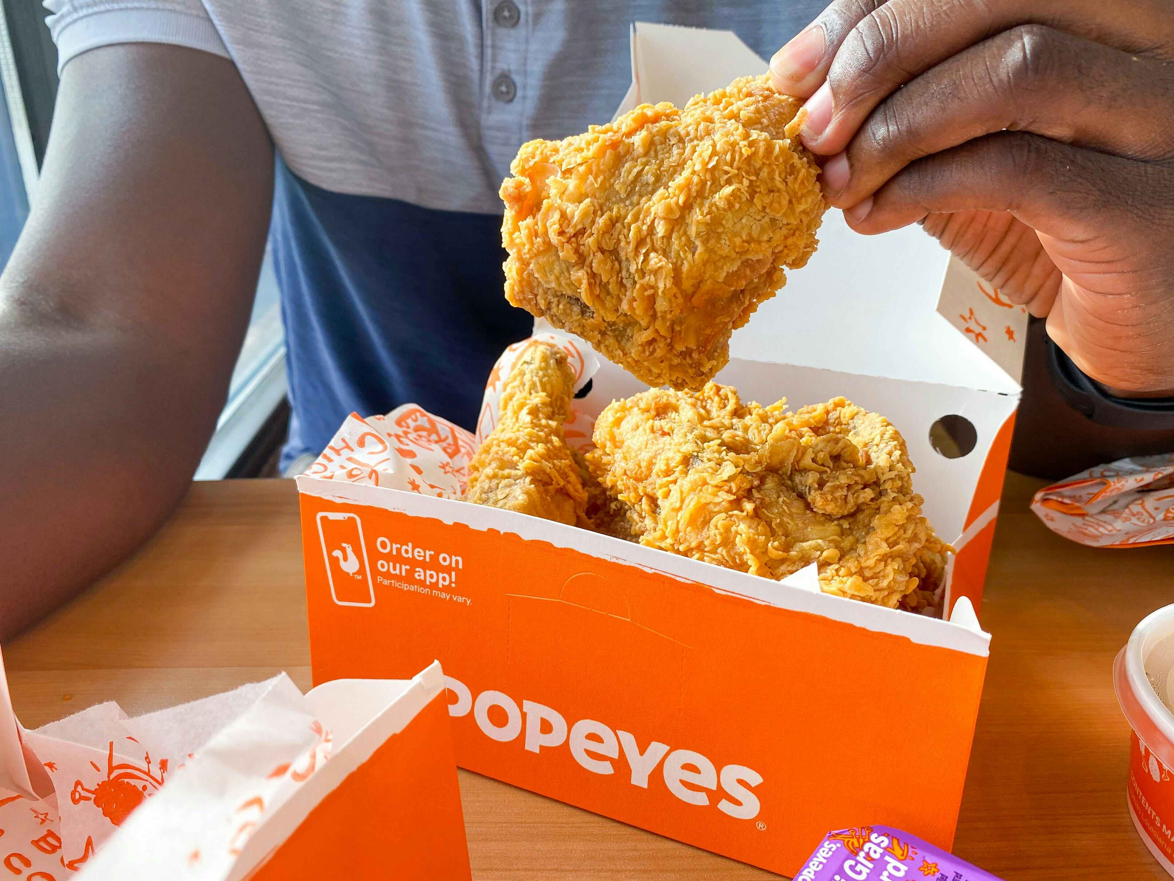 A man taking a piece of chicken out of a box of Popeyes chicken.