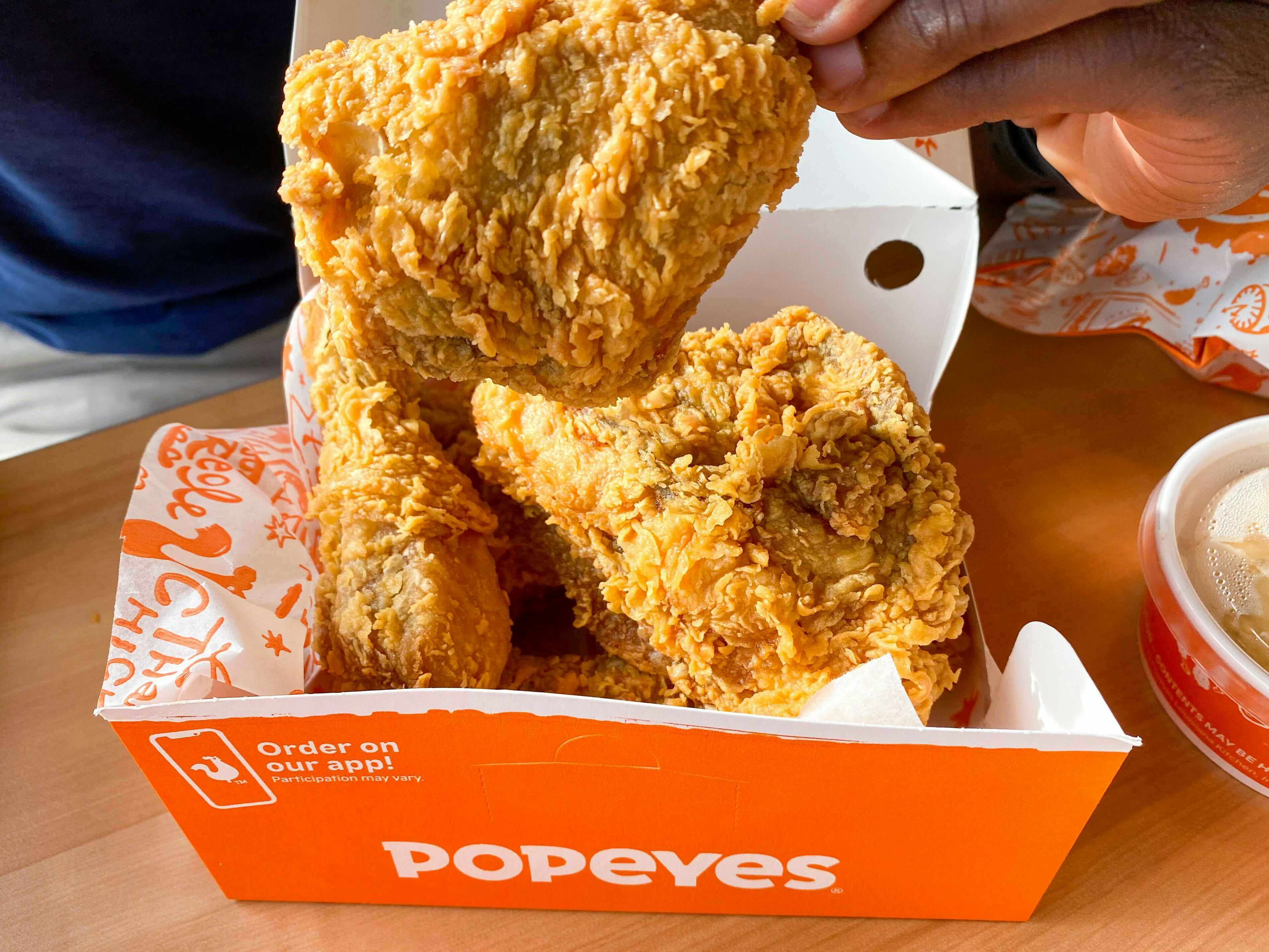 A person's hand taking a piece of chicken out of a box of Popeyes chicken sitting on a table.