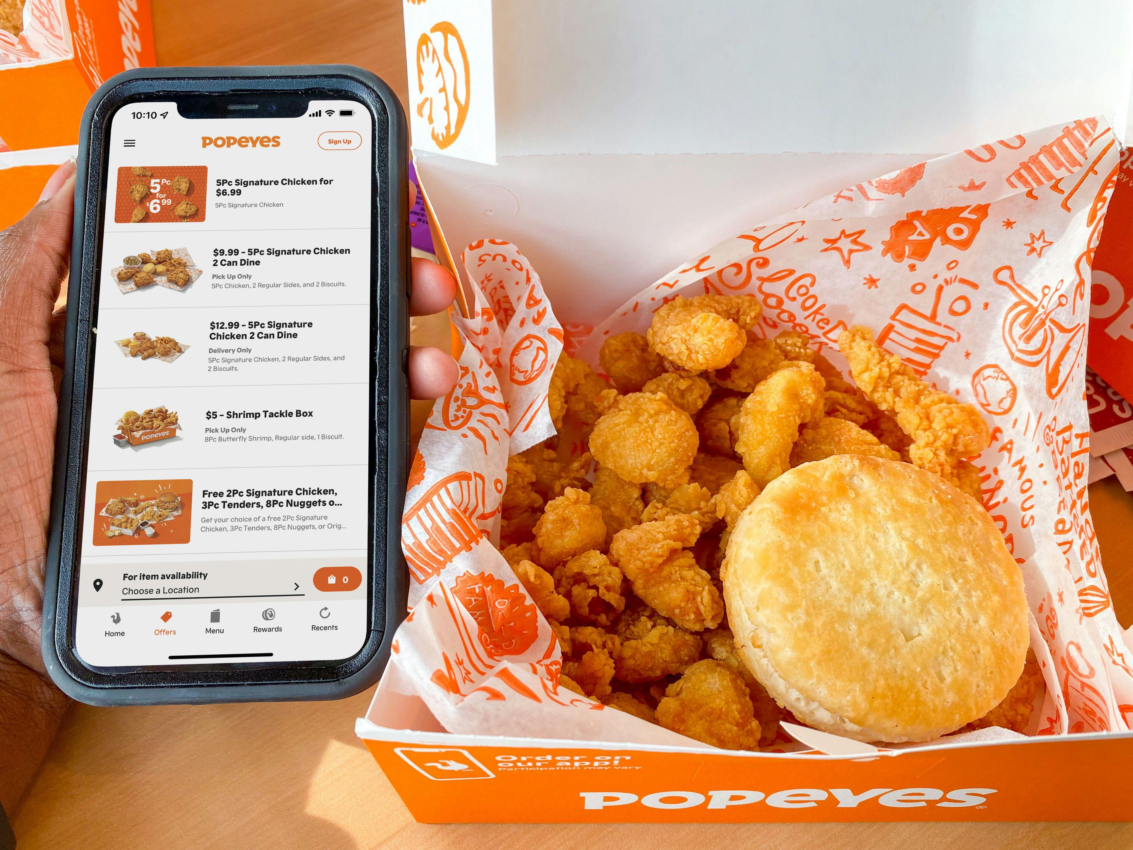 A person holding an iPhone displayng the Popeyes app next to a box of Popeyes chicken and a biscuit sitting on a table.