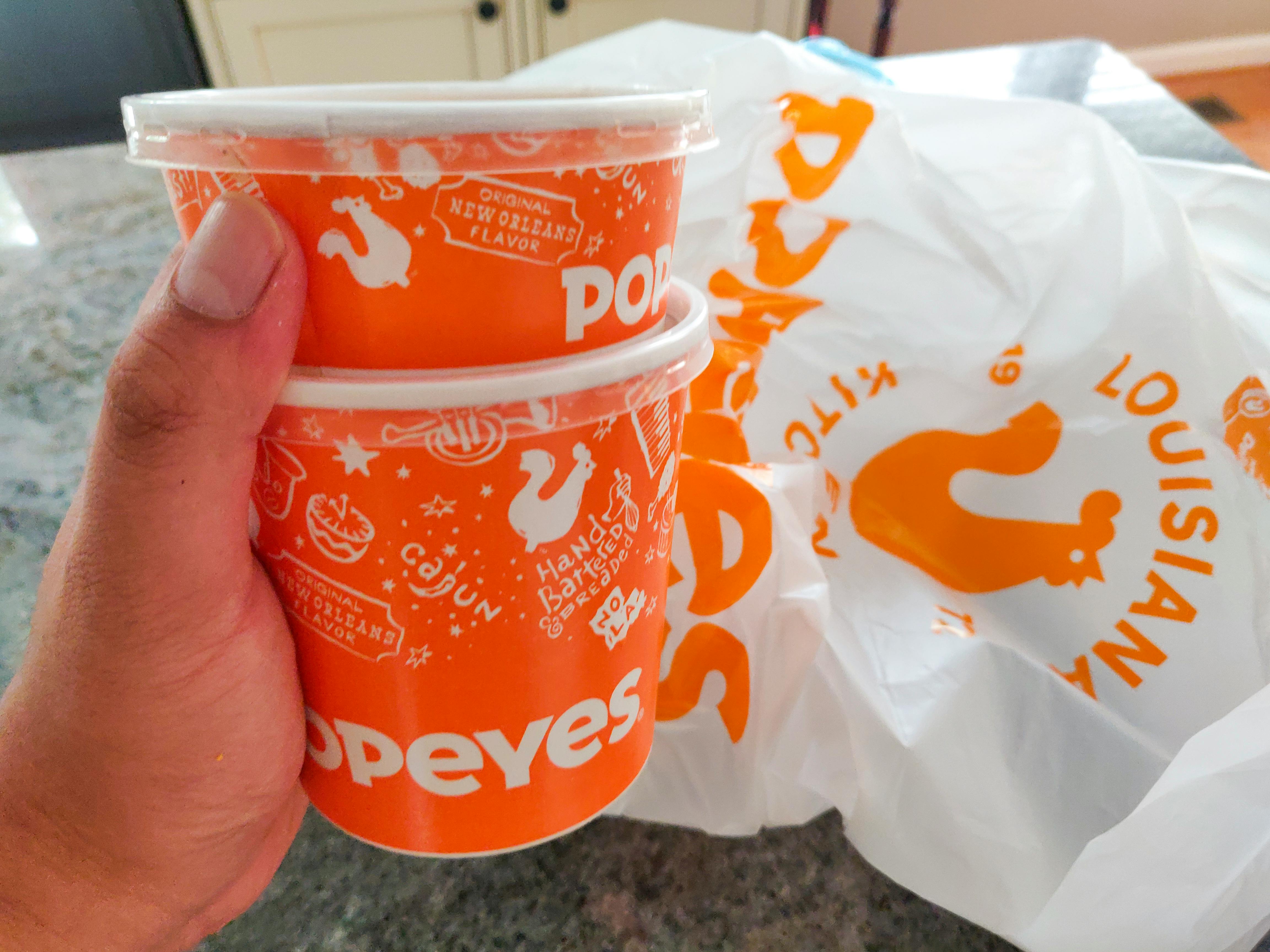 A person's hand holding two containers of Popeyes side dishes next to a Popeyes takeout bag on a counter.