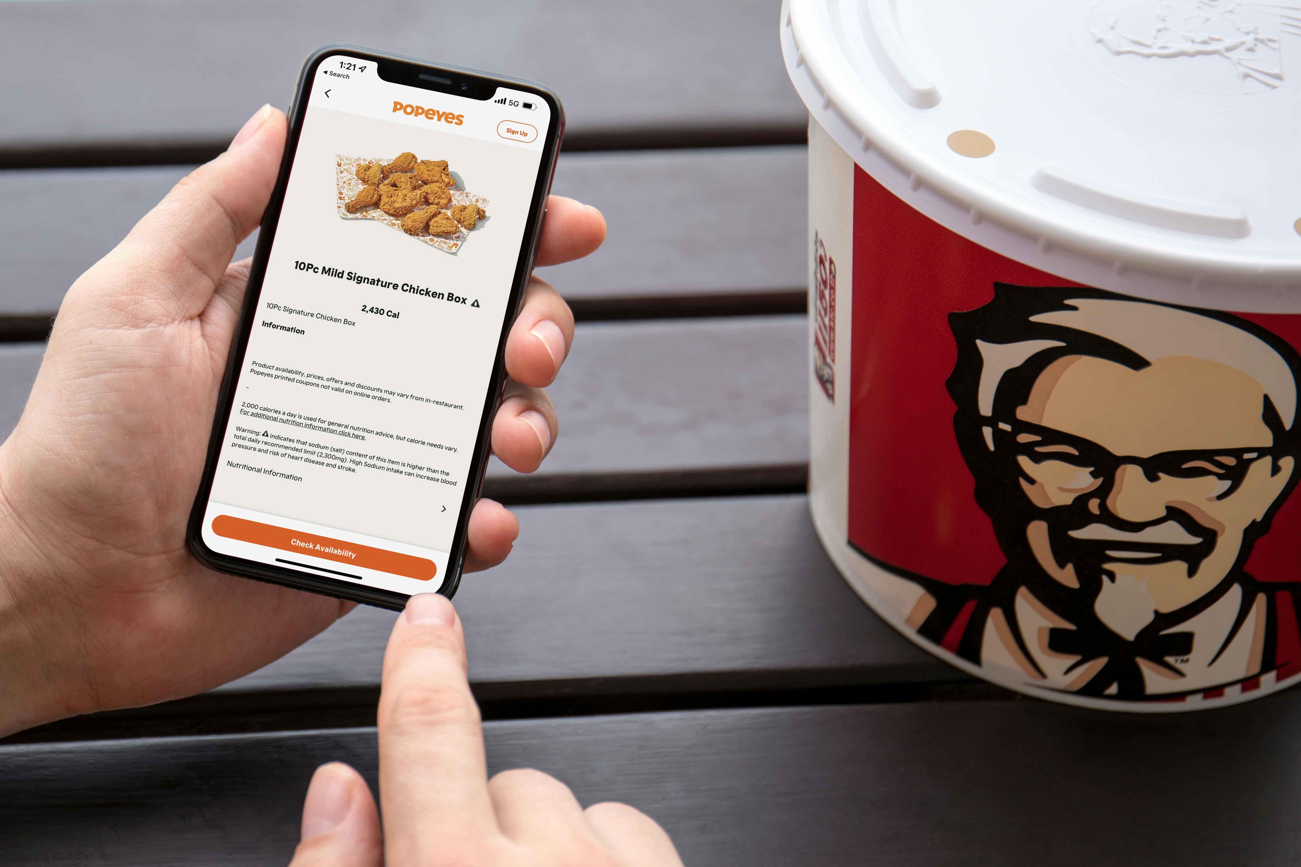 A person's hand holding an iPhone displaying the Popeyes app next to a KFC chicken bucket.