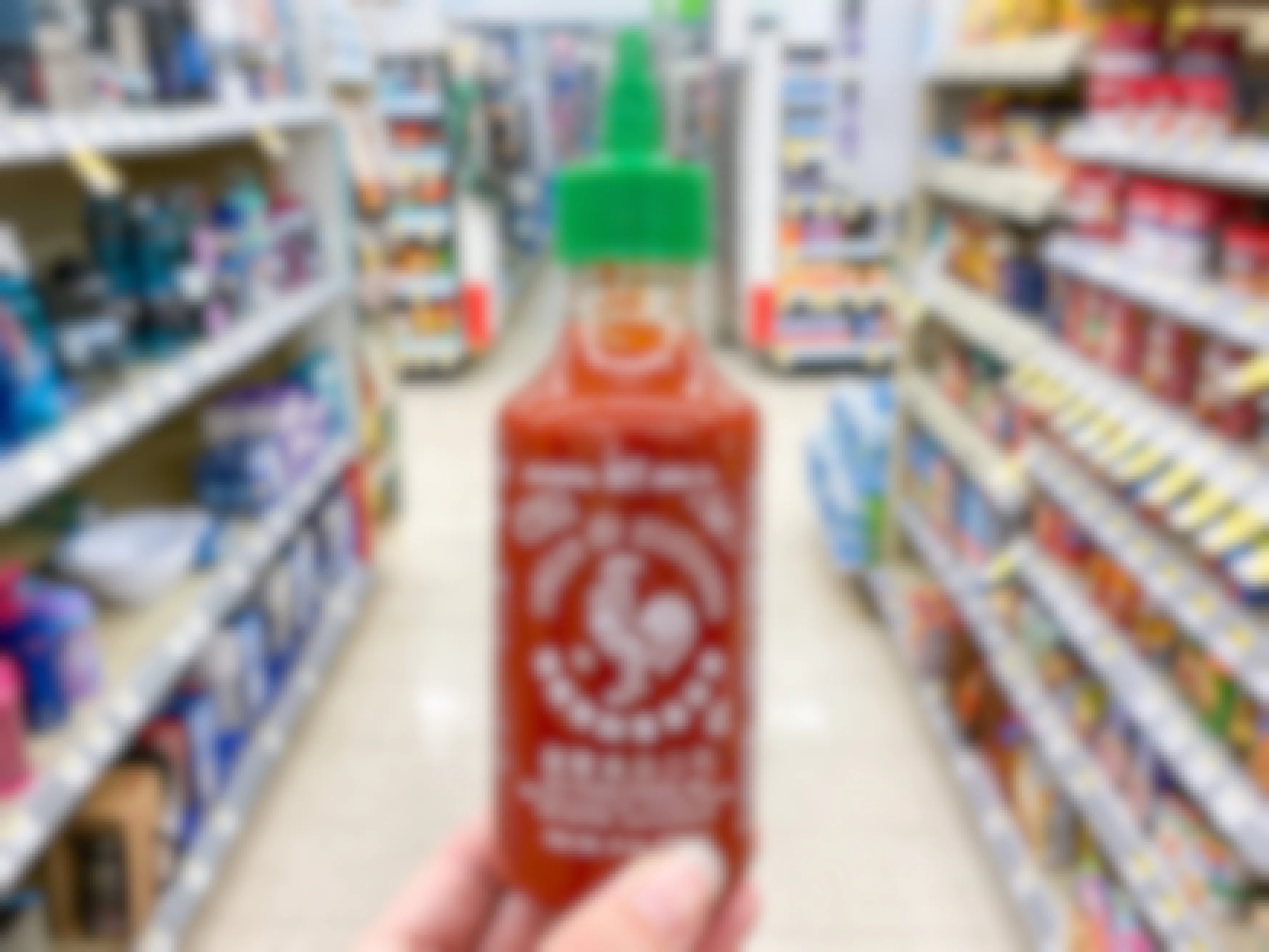 A person's hand holding up a bottle of Sriracha in an aisle at Walgreens.