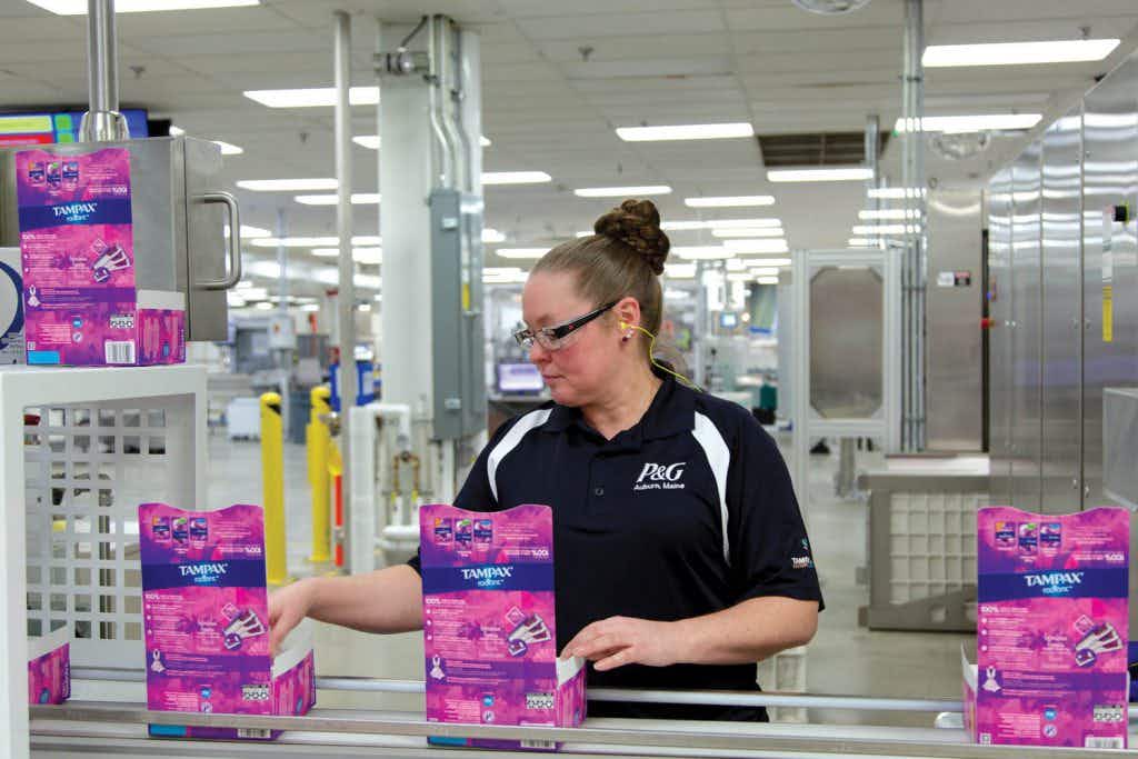 A woman wearing a P&G t-shirt, standing behind a conveyer belt with Tampax tampon boxes running along it.