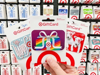 Three Target gift cards held in a person's hand in front of the wall of gift cards at Target.
