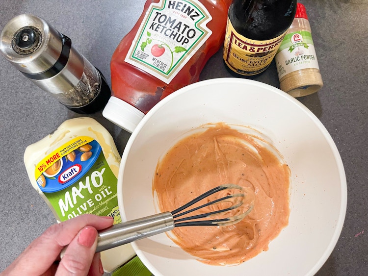 A mixing bowl with homemade sauce with mayo, black pepper, ketchup, Worcestershire sauce, and garlic powder, with a person whisking it 