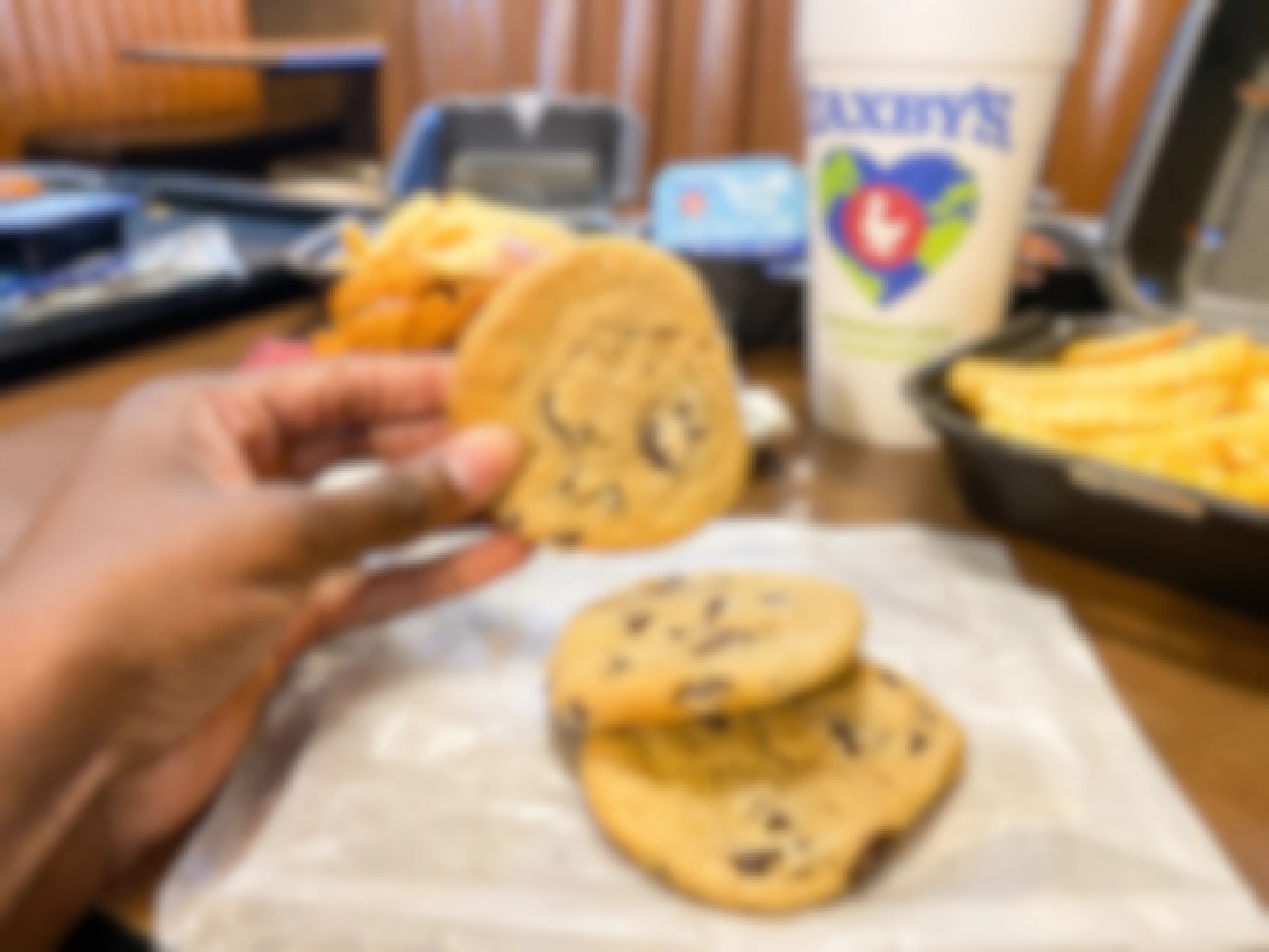 A person's hand holding a chocolate chip cookie above two more cookies on a table at Zaxby's.