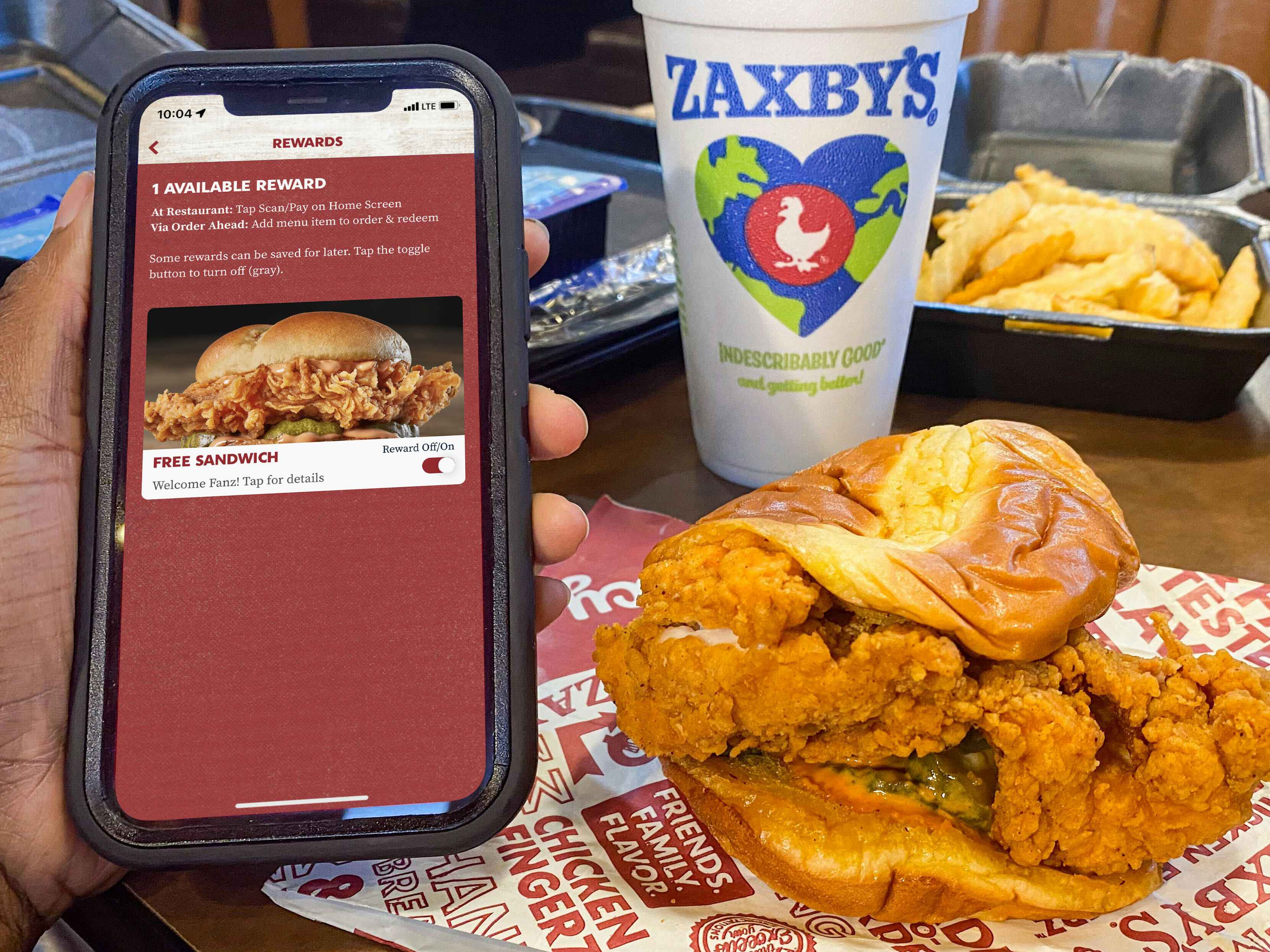 A person's hand holding a cell phone displaying the Rewards page of the Zaxby's app next to a chicken sandwich, a drink, and a box of fries on a table at Zaxby's.