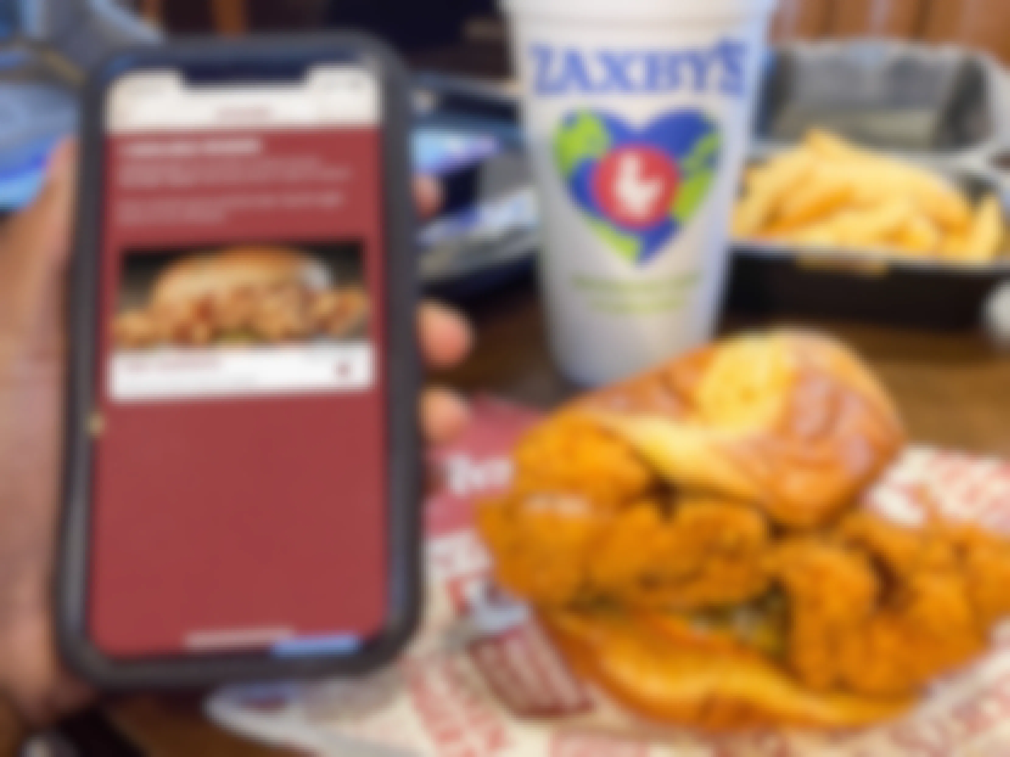A person's hand holding a cell phone displaying the Rewards page of the Zaxby's app next to a chicken sandwich, a drink, and a box of fries on a table at Zaxby's.