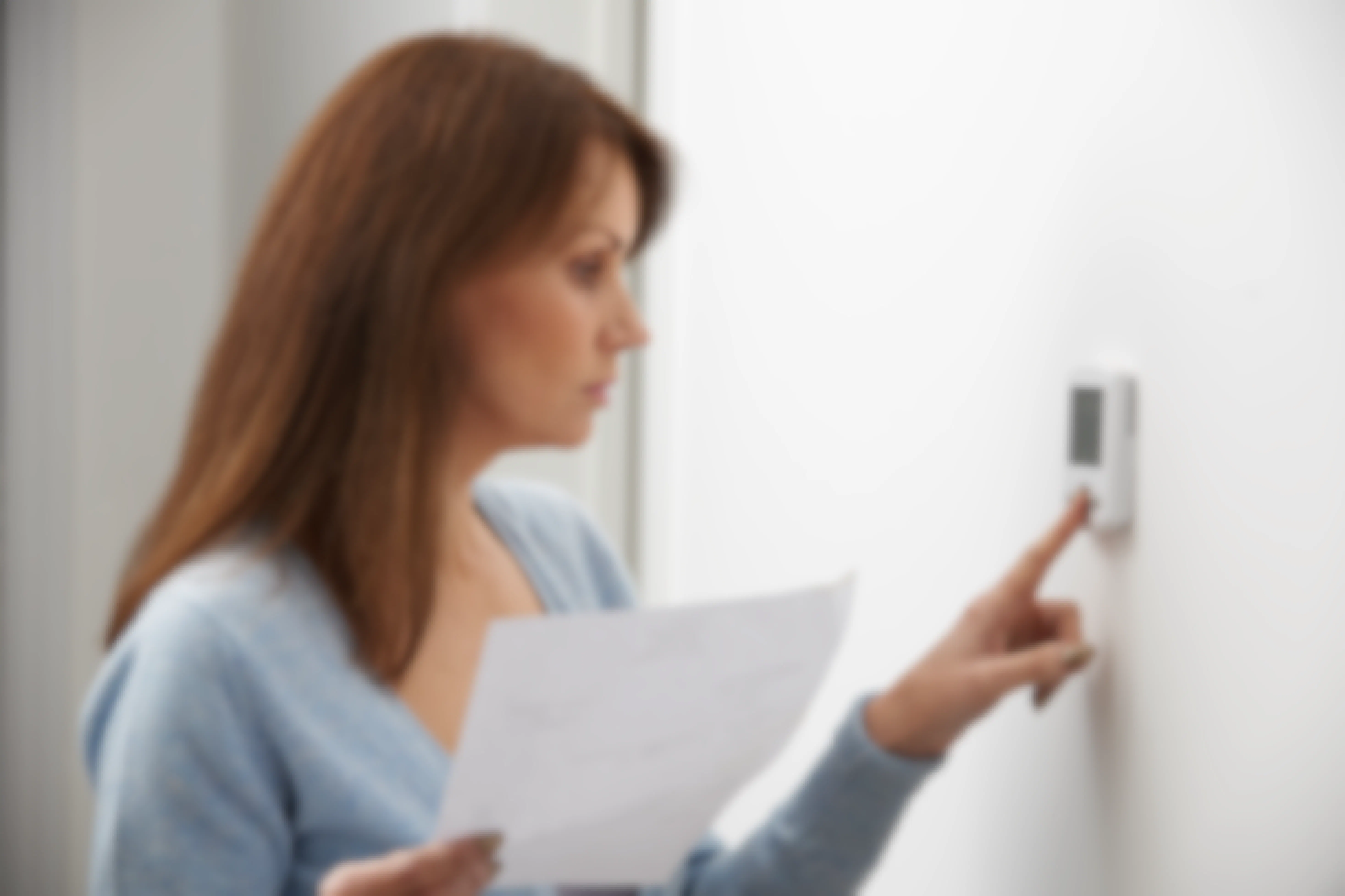 A woman holding an energy bill and changing the temperature on her thermostat on the wall.