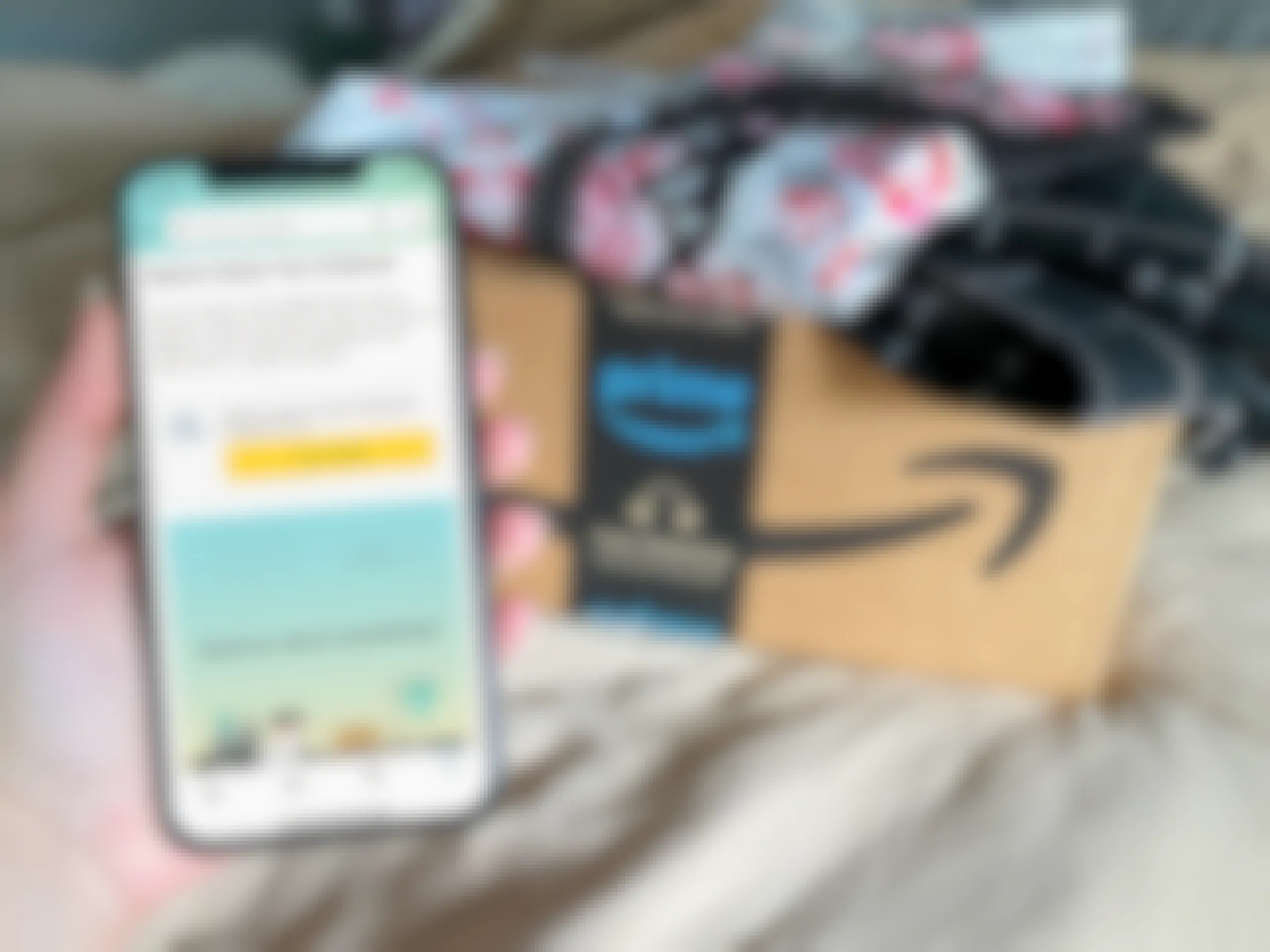 A person's hand holding an iPhone displaying the "Return Items You Ordered" page on the Amazon app next to an Amazon Prime box with clothes on top.
