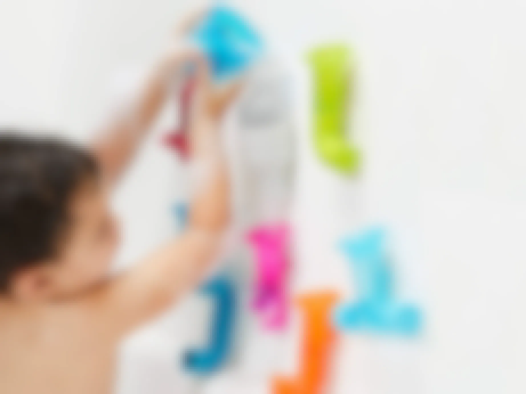 A child in a bath, pouring water into some Boon bath toys suction cupped to the wall.