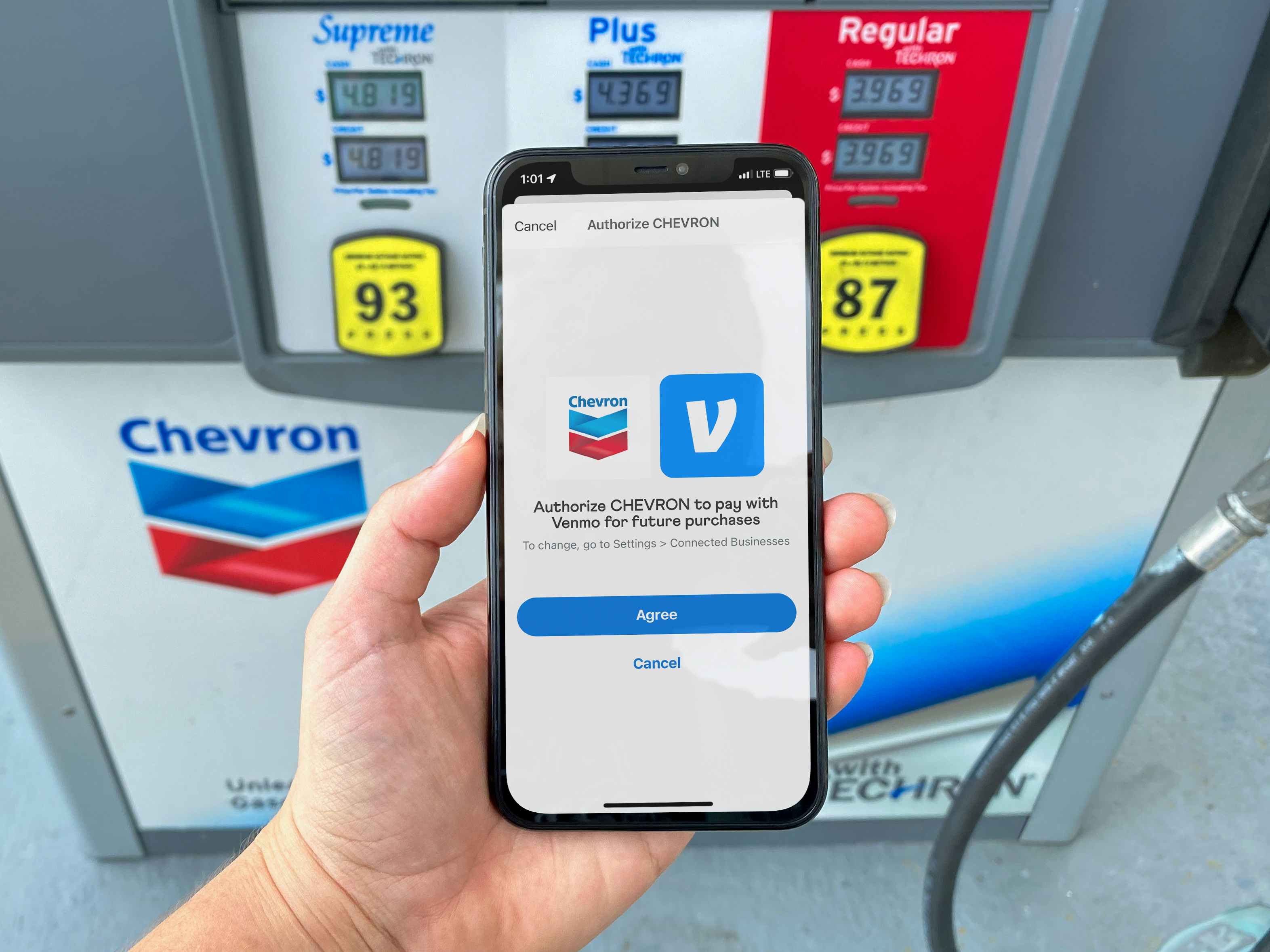 A person's hand holding an iPhone displaying the venmo authorization page in the Chevron mobile app in front of a Chevron gas station pump.