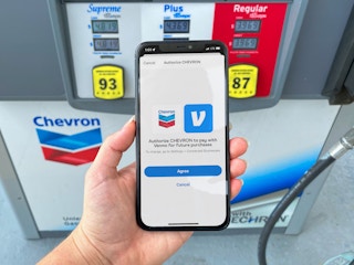 A person's hand holding an iPhone displaying the venmo authorization page in the Chevron mobile app in front of a Chevron gas station pump.