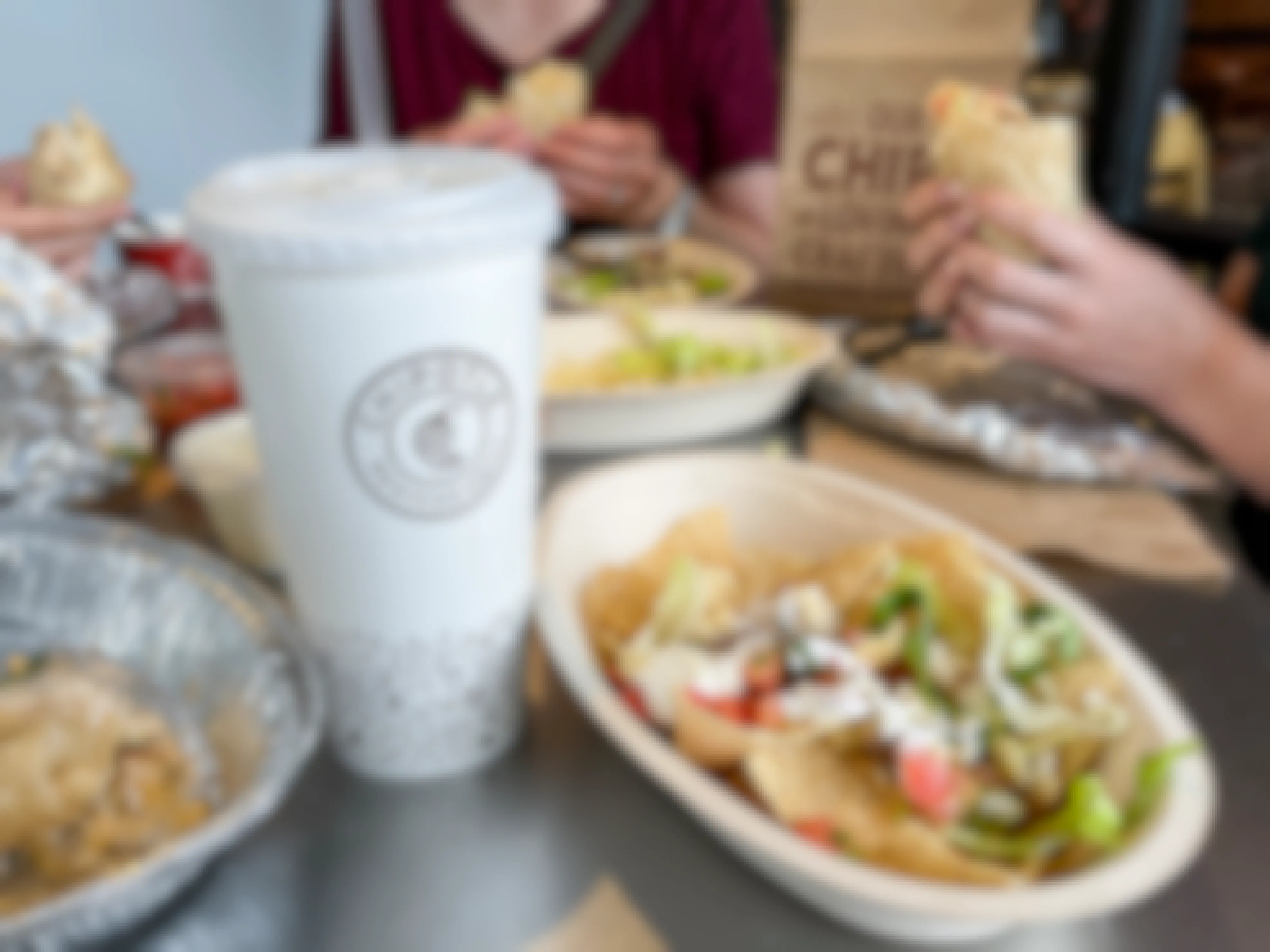 Three people holding burittos at a table filled with food. A drink cup is in the for front clearly displaying the Chipotle logo.