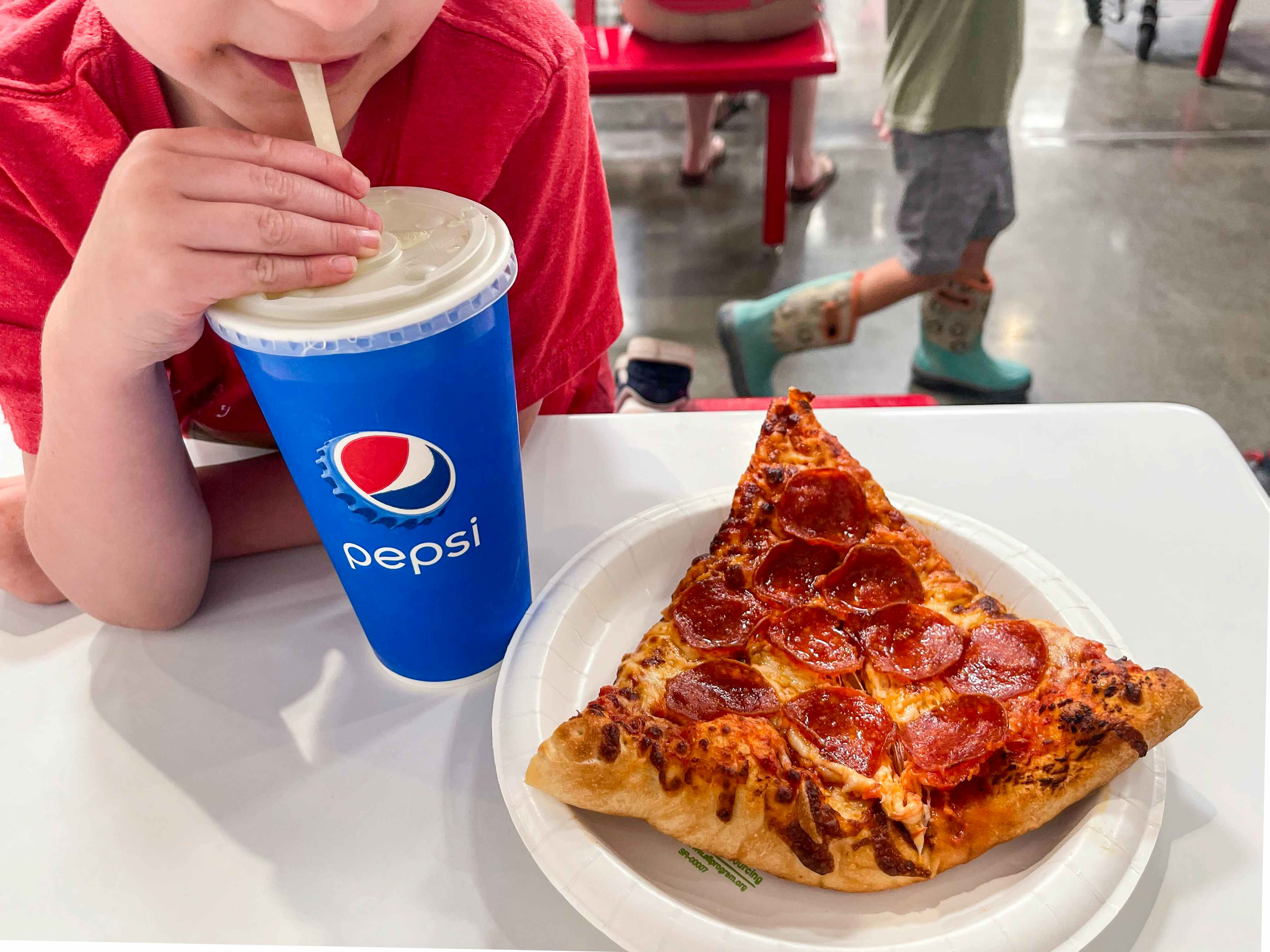 A child drinking out of a disposable Pepsi drink cup with a piece of pepperoni pizza on a plate next to him in the Costco food court.