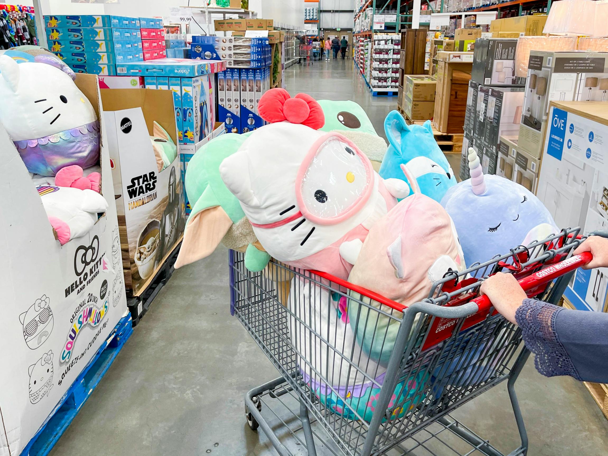 A person pushing a Costco shopping cart filled with big Sqishmallows next to some Squishmallow display boxes.