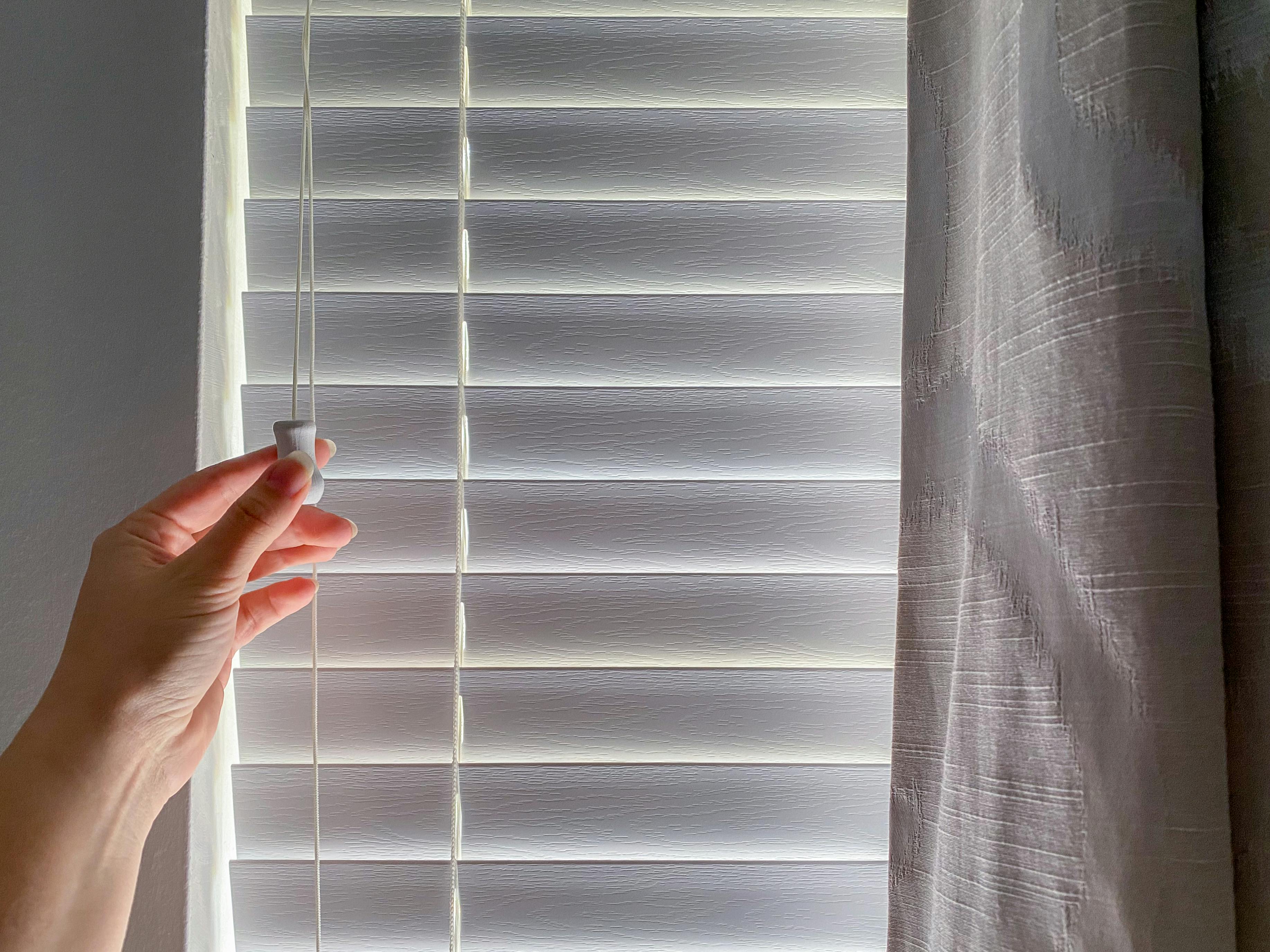 A person's hand closing the blinds of a window where the sun is shining through.