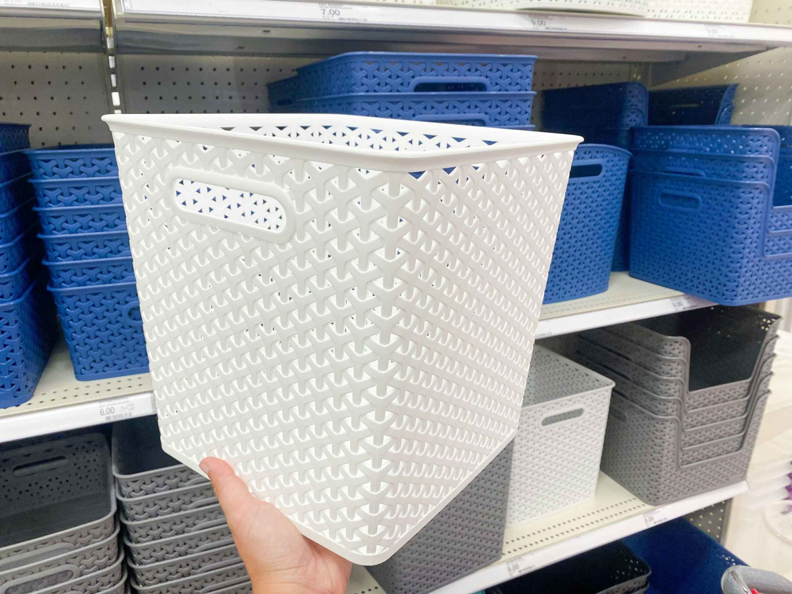 A hand holding a white decorative storage basket in front of a shelf with other storage baskets