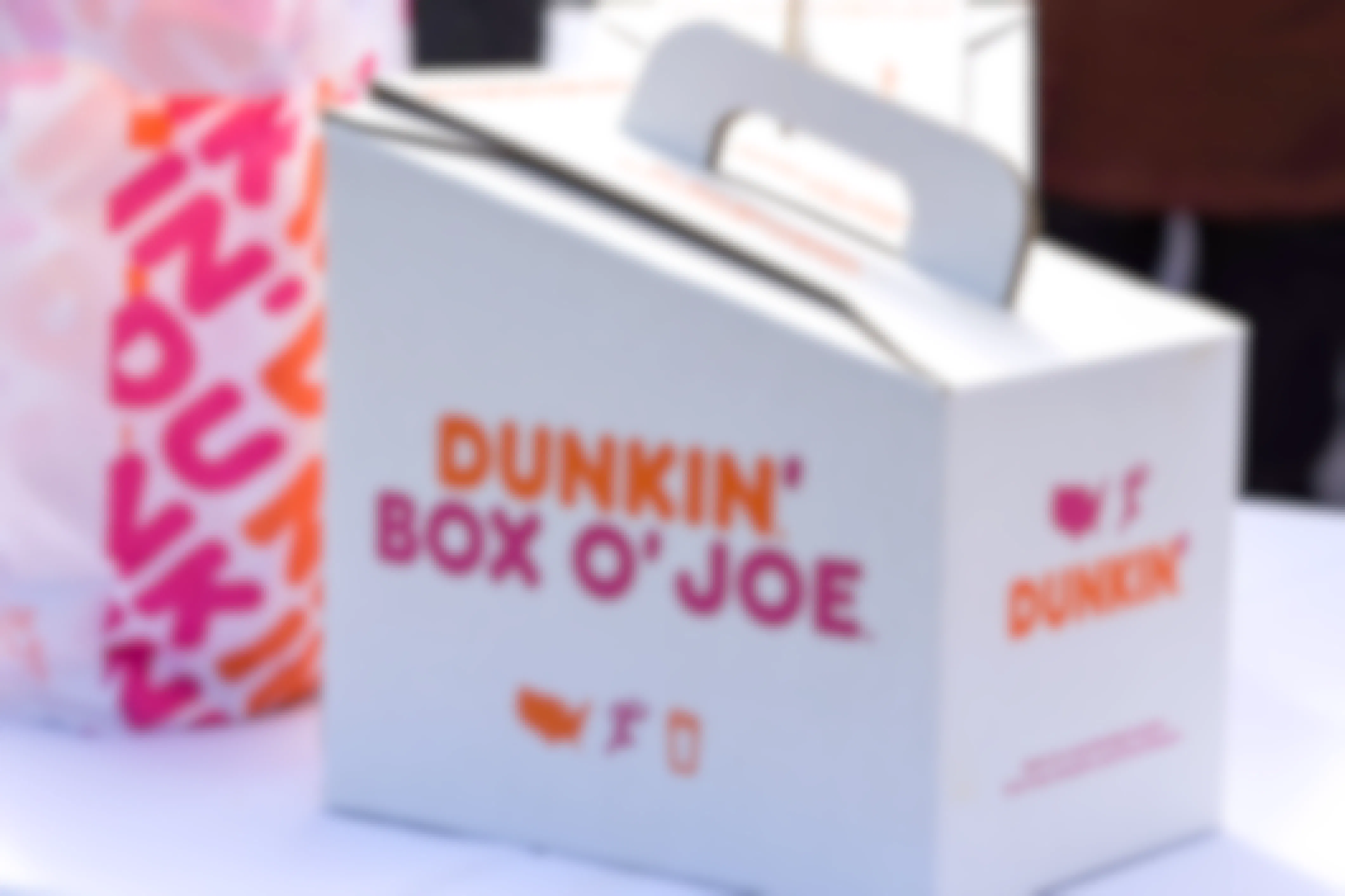 A Dunkin Donuts coffee Box O' Joe on a table next to some Dunkin to-go bags.