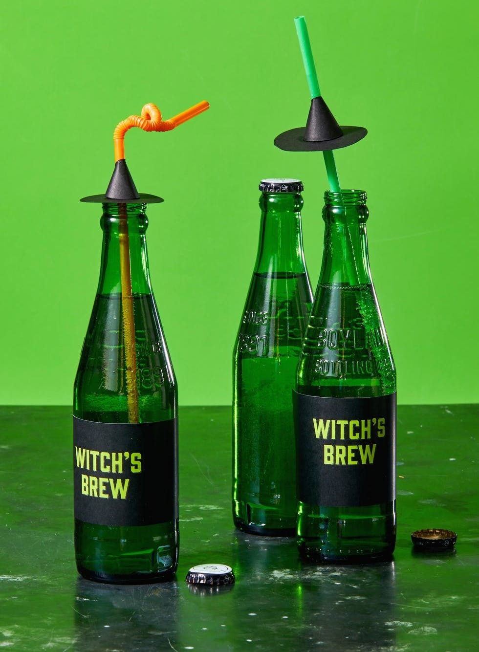 Three green glass bottles wrapped in black Witch's Brew labels sitting on a green table with a green background.