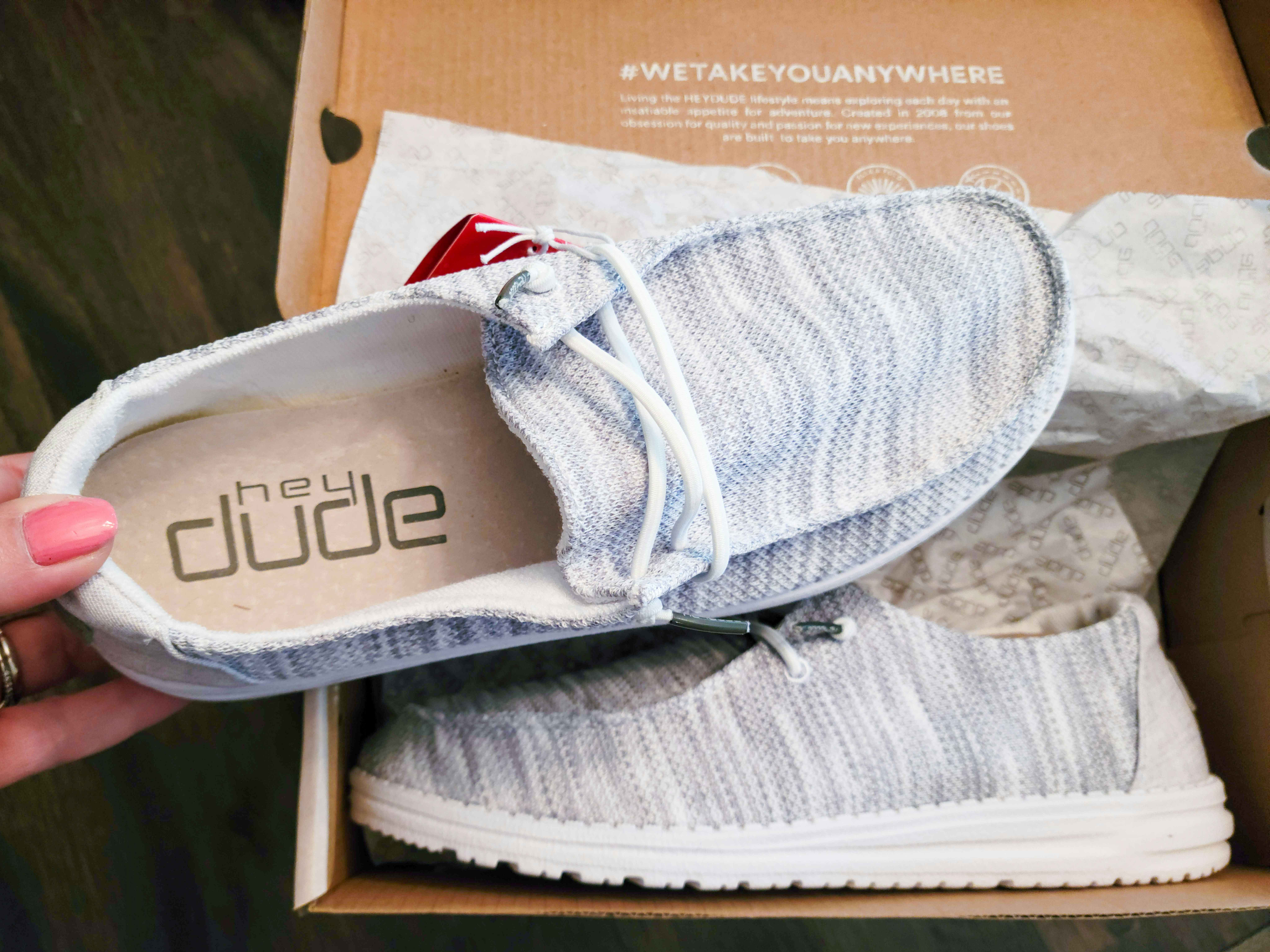 A person's hand holding a grey Hey Dude Sneaker, showing off the inner label above the other shoe still in the box.