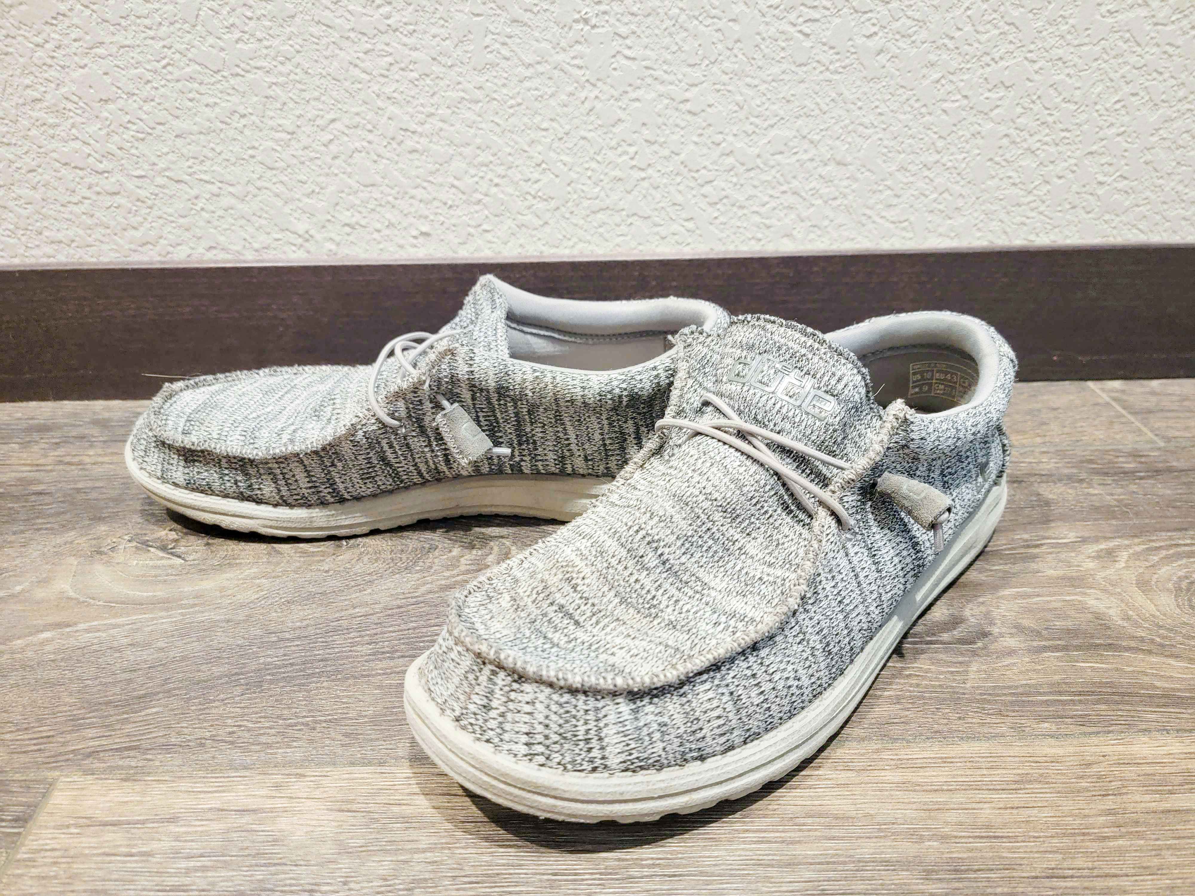 A pair of Hey Dude shoes on a wood floor next to a wall in someone's house.