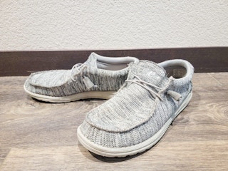 A pair of Hey Dude shoes on a wood floor next to a wall in someone's house.