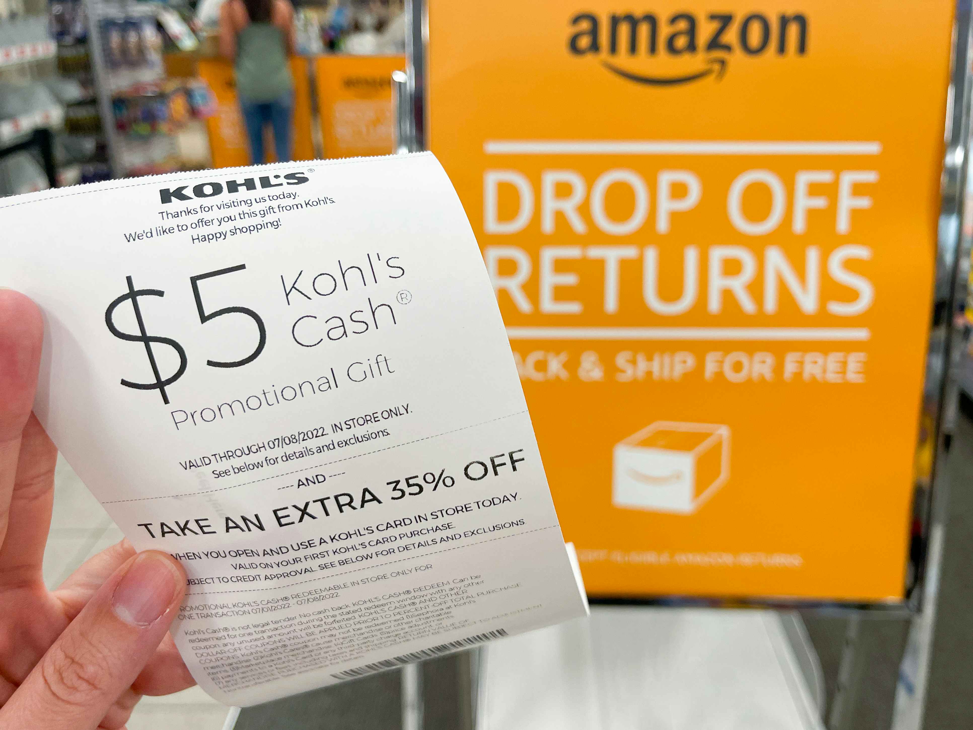 A person's hand holding a $5 Kohl's Cash coupon on a receipt for returning an Amazon package next to the Amazon drop off returns sign inside Kohl's.
