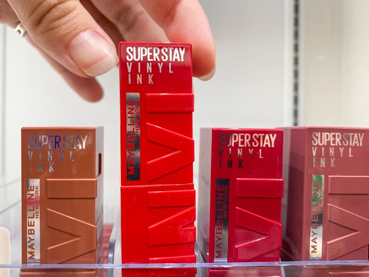 A person's hand reaching to take a Maybelline Super Stay Vinyl lipstick from a display in a store.