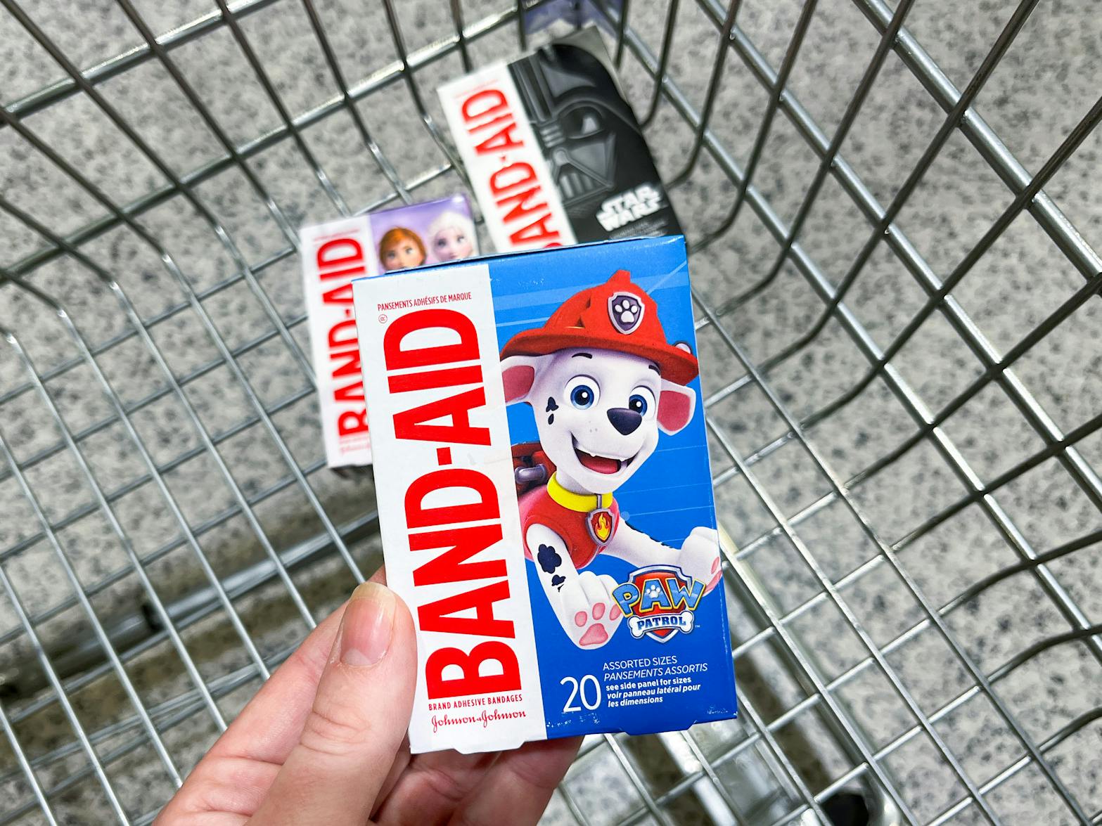 A person's hand holding a box of Paw Patrol themed Band-Aids above some Star Wars and Frozen themed Band-Aids in a shopping cart.