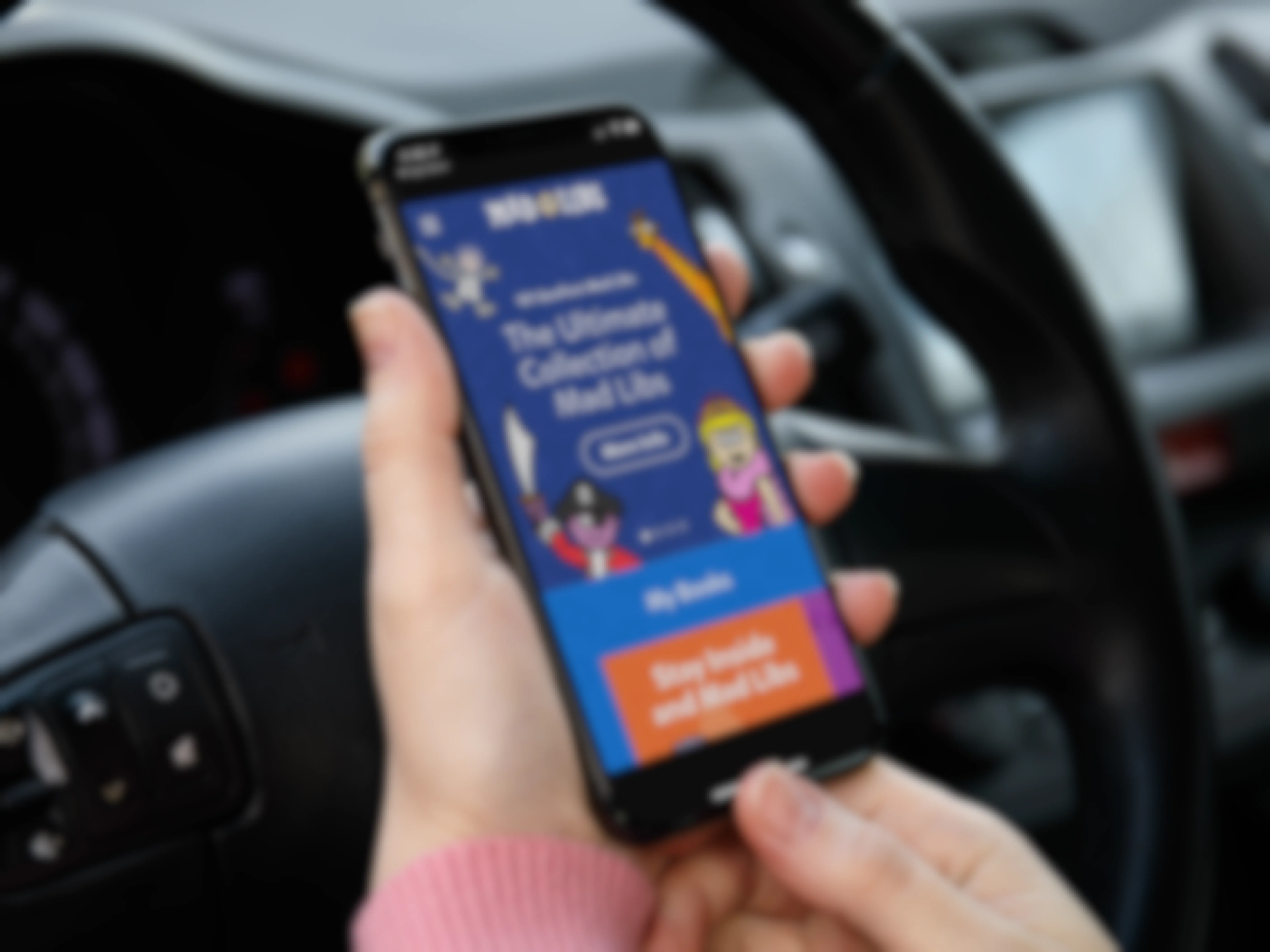 A person sitting in a car, holding an iPhone displaying the Mad Libs app home screen.