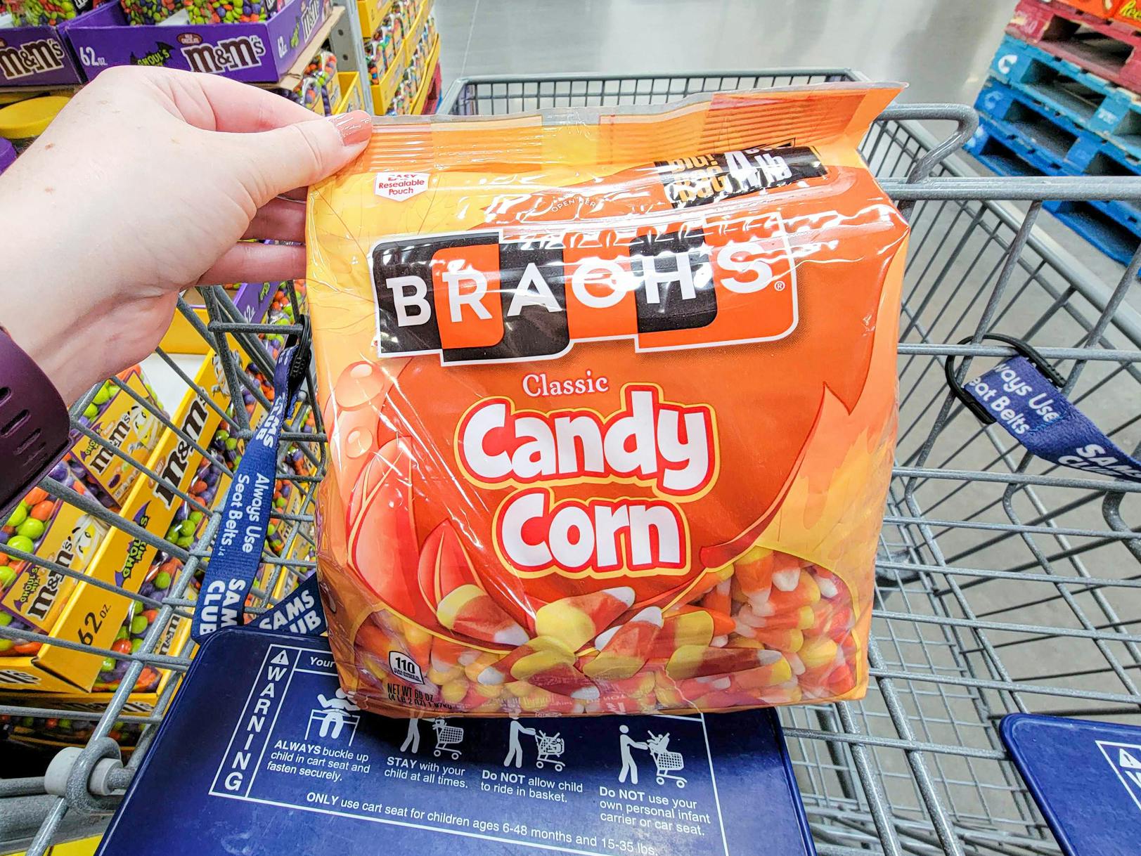A person's hand putting a large bag of candy corn in a cart at Sam's Club.