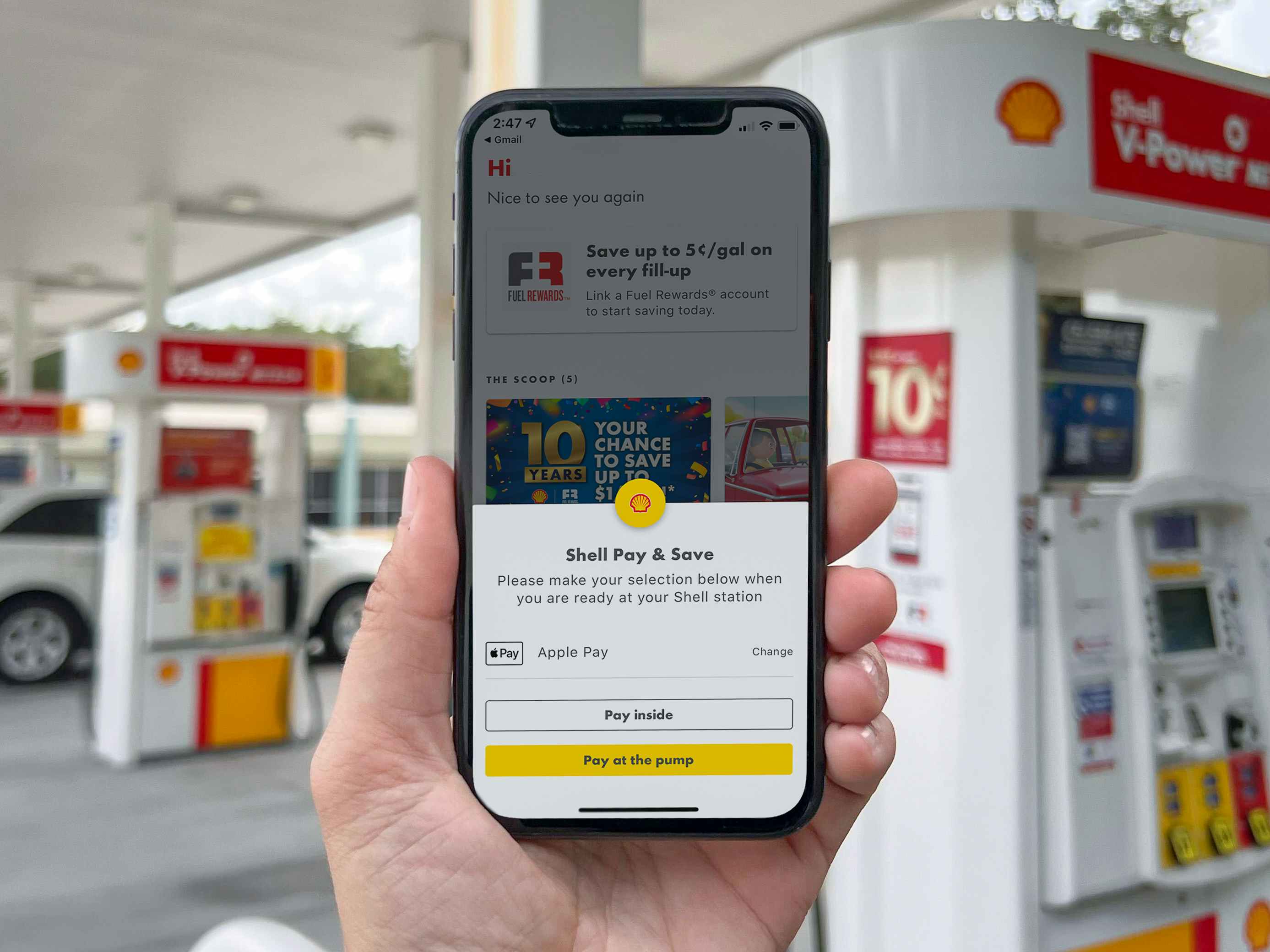 A person holding up a cell phone displaying the Shell Pay & Save page on the Shell rewards app in front of a gas pump at a Shell gas station.