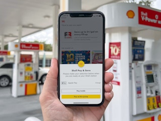 A person holding up a cell phone displaying the Shell Pay & Save page on the Shell rewards app in front of a gas pump at a Shell gas station.